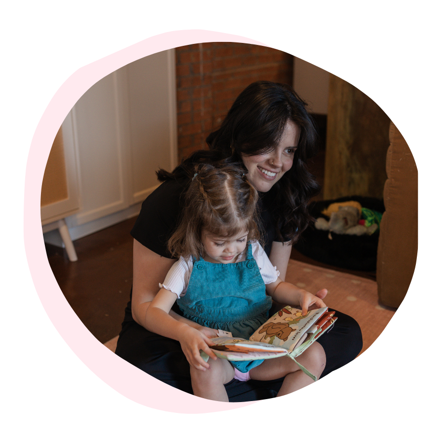 A woman and a young girl sitting together on the floor, reading a picture book, with the girl sitting on the woman's lap.