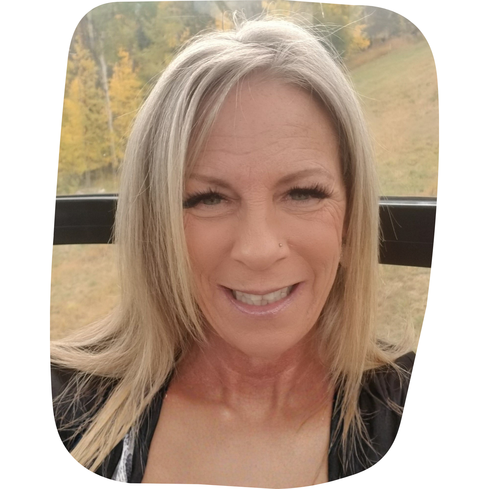 A smiling woman with blonde hair, wearing false eyelashes and a nose piercing. She is outdoors with trees in the background.