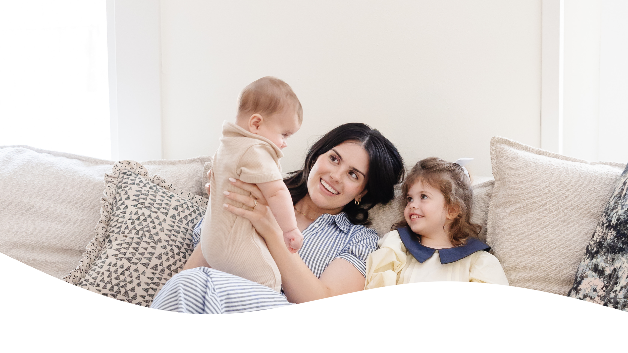 Woman and two young girls sitting on a sofa, smiling and playing together in a bright living room.