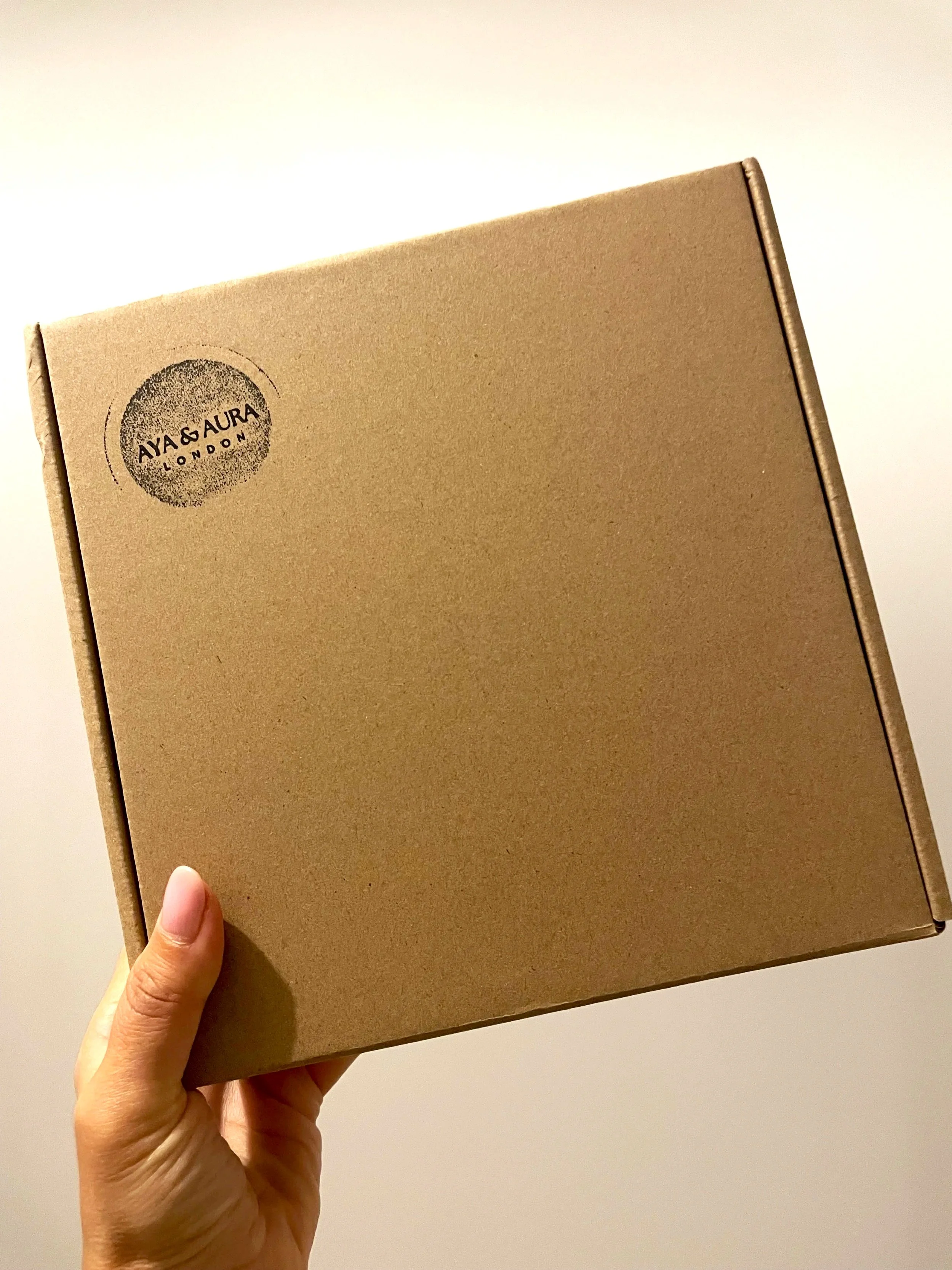 A hand holding a brown cardboard parcel box with a round stamped logo that says 'AYA & AURA LONDON' in the top left corner.