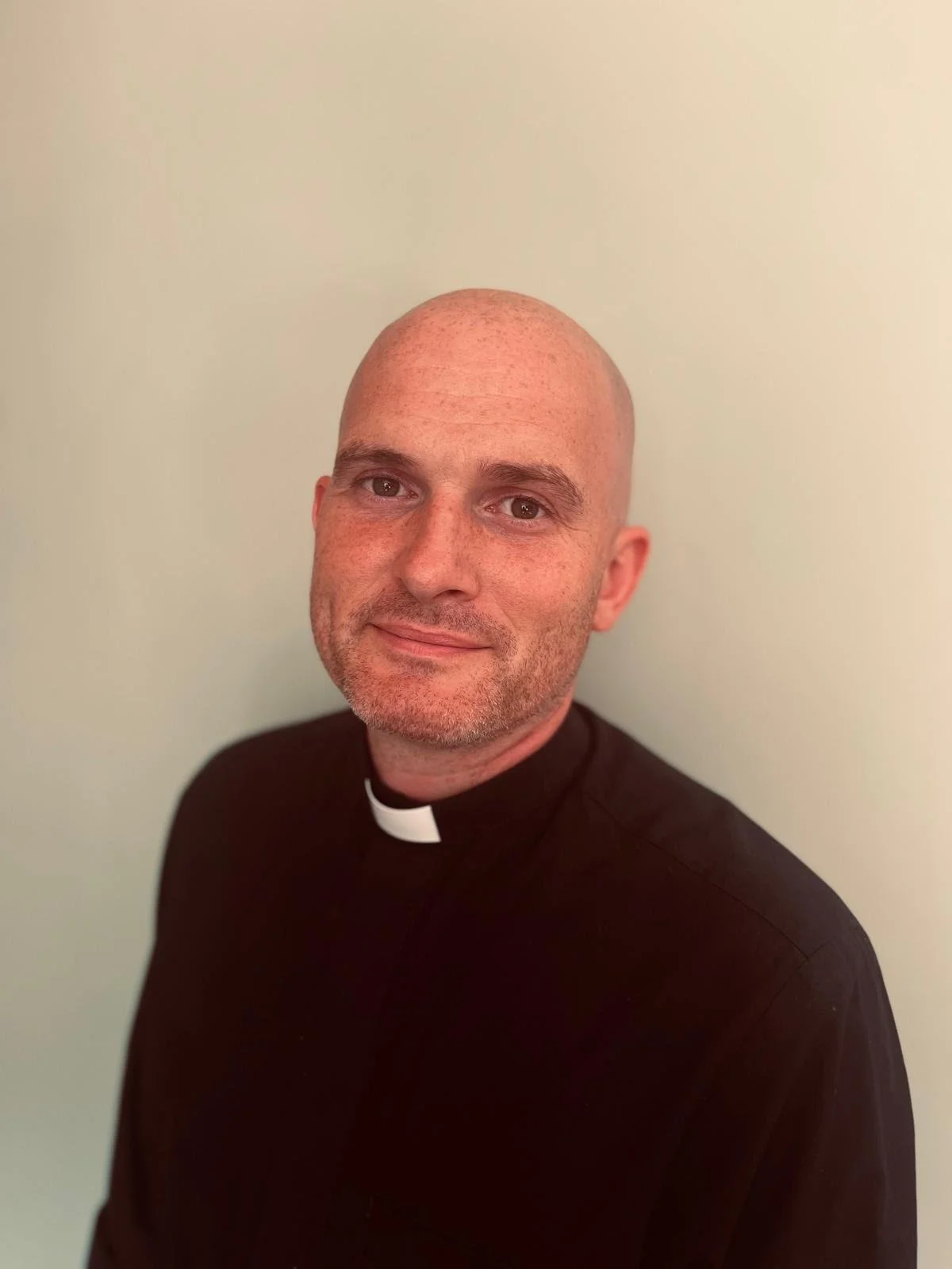 A man with a shaved head and a short beard, wearing a black clergy shirt with a white collar, standing against a plain light-colored wall