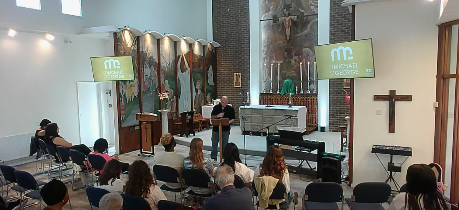 A church interior with a congregation seated facing an altar. A speaker is at a lectern reading from a book. The altar features candles, a crucifix on the wall, and a religious painting backdrop. Two large screens display the text "ST. MICHAEL ST. GEORGE."