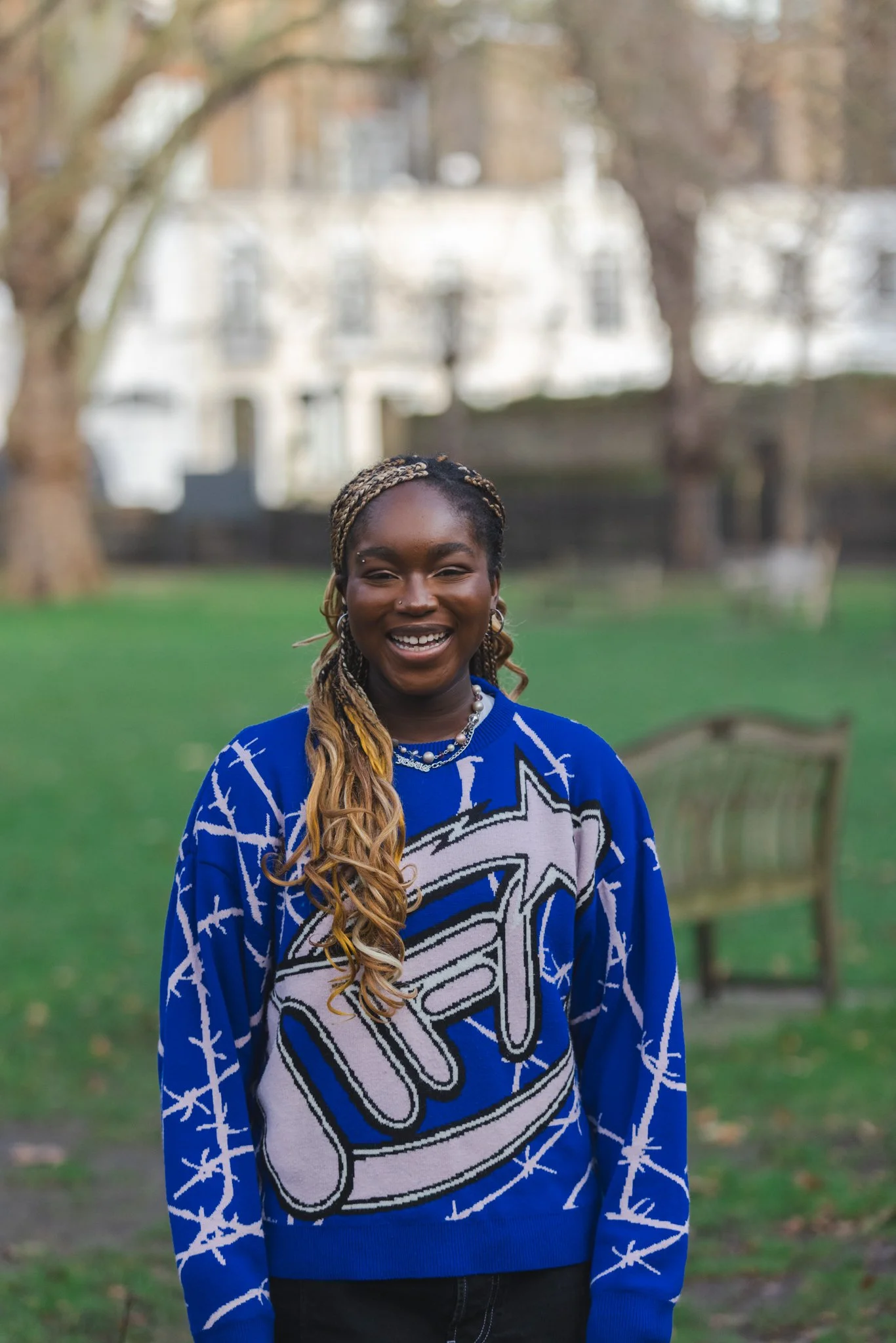 A young woman with braided hair smiling and standing outdoors in a park. She is wearing a blue sweater with large graphic text and barbed wire patterns, along with jewelry including earrings, a necklace, and a nose ring. In the background, there are green trees, a wooden bench, and a blurred white building.