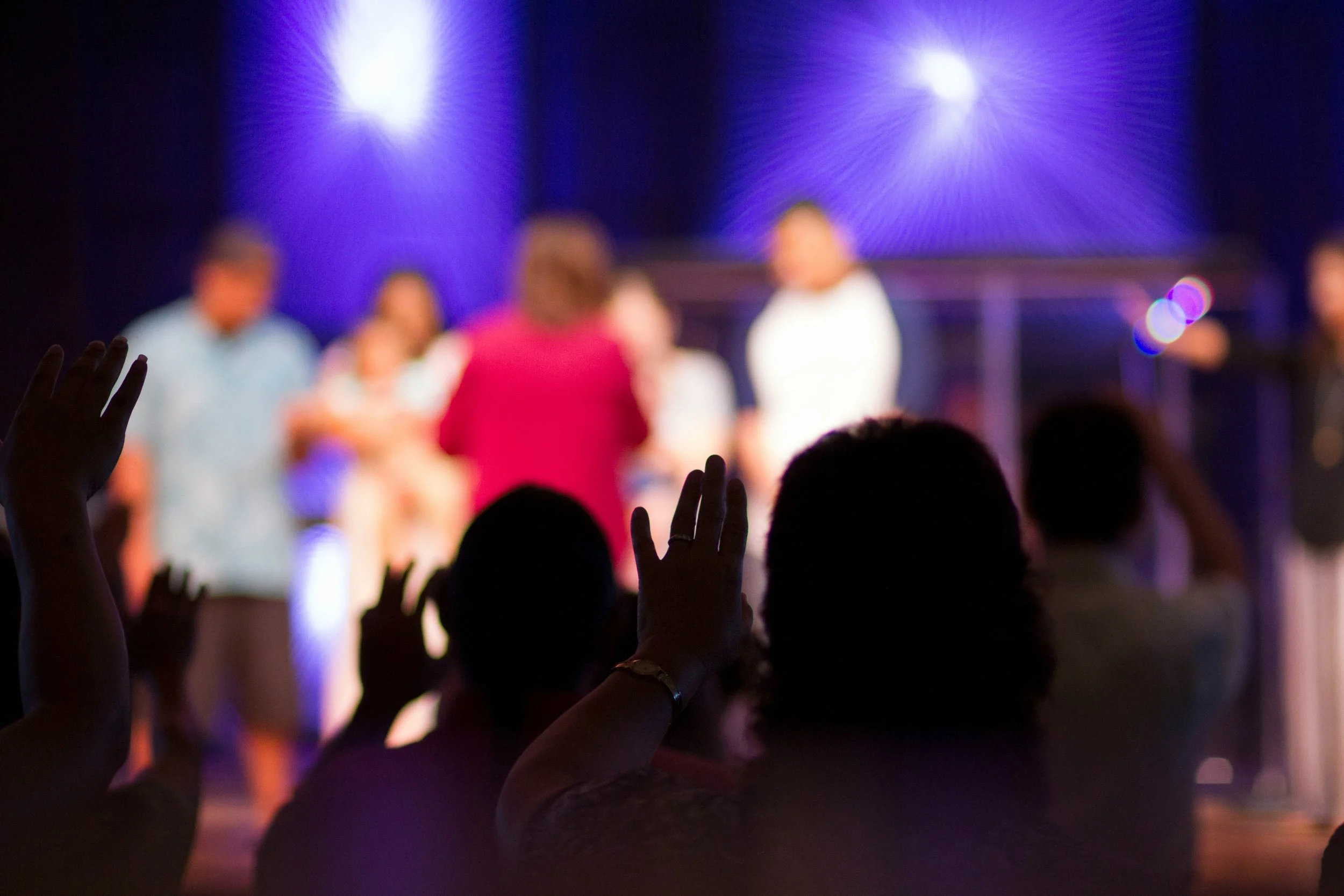 People in a dark room raising their hands while standing in front of a stage with a blue and purple light display.