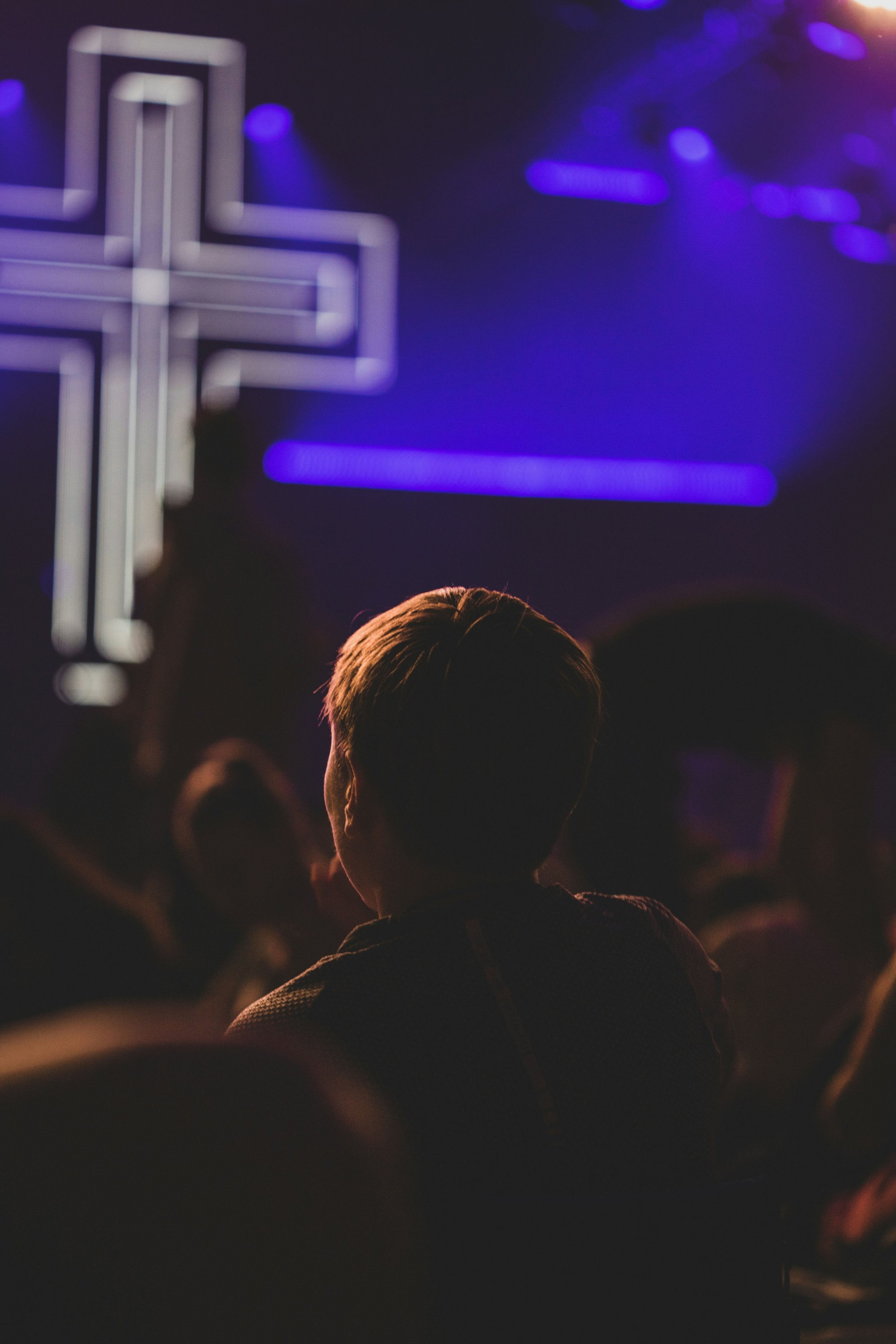 A person with light hair sitting in a dark room, facing a brightly lit cross on a stage, with purple and blue stage lights in the background.