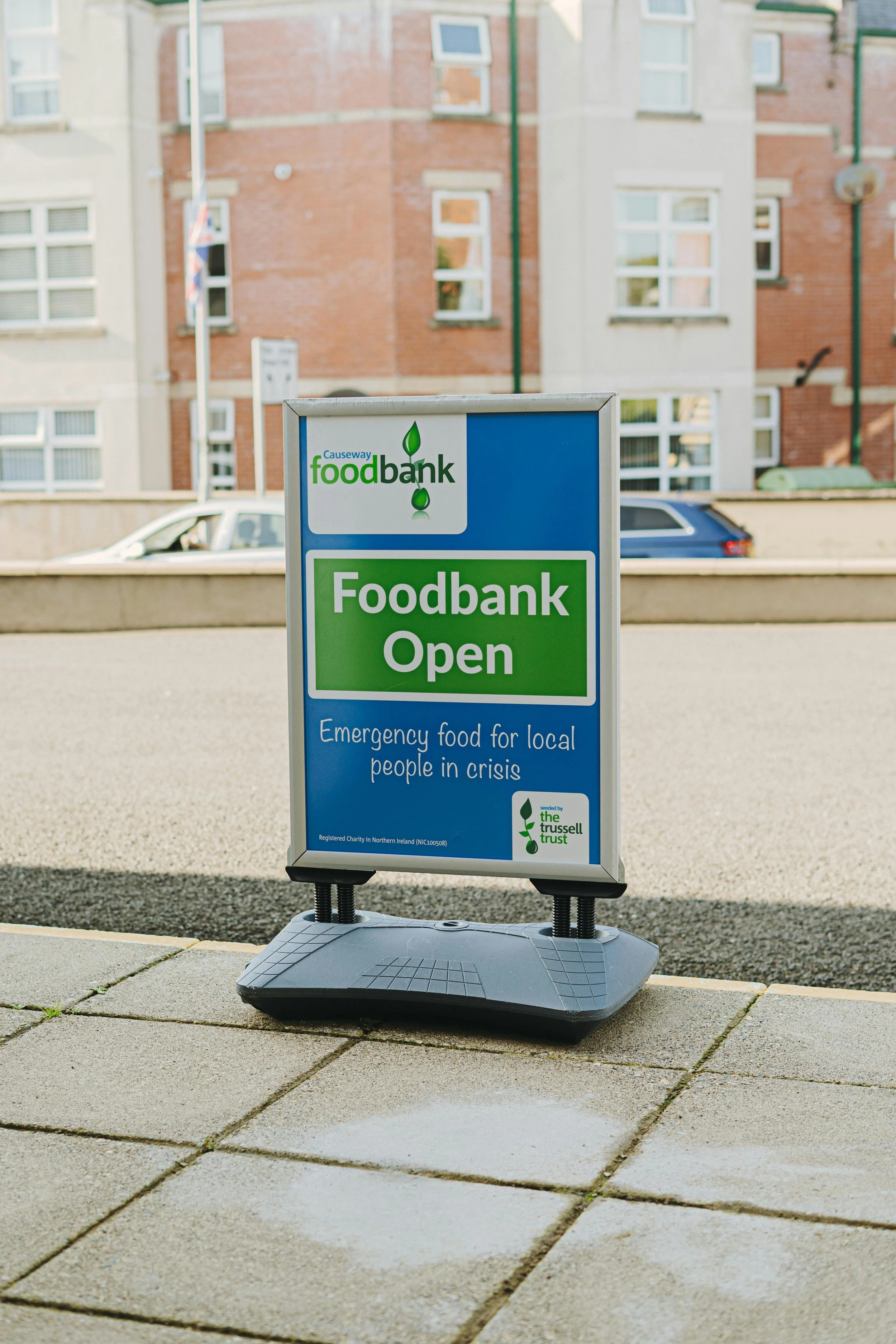 A sign for a food bank, indicating it is open and providing emergency food for local people in crisis, placed on a sidewalk in an urban area with buildings and cars in the background.