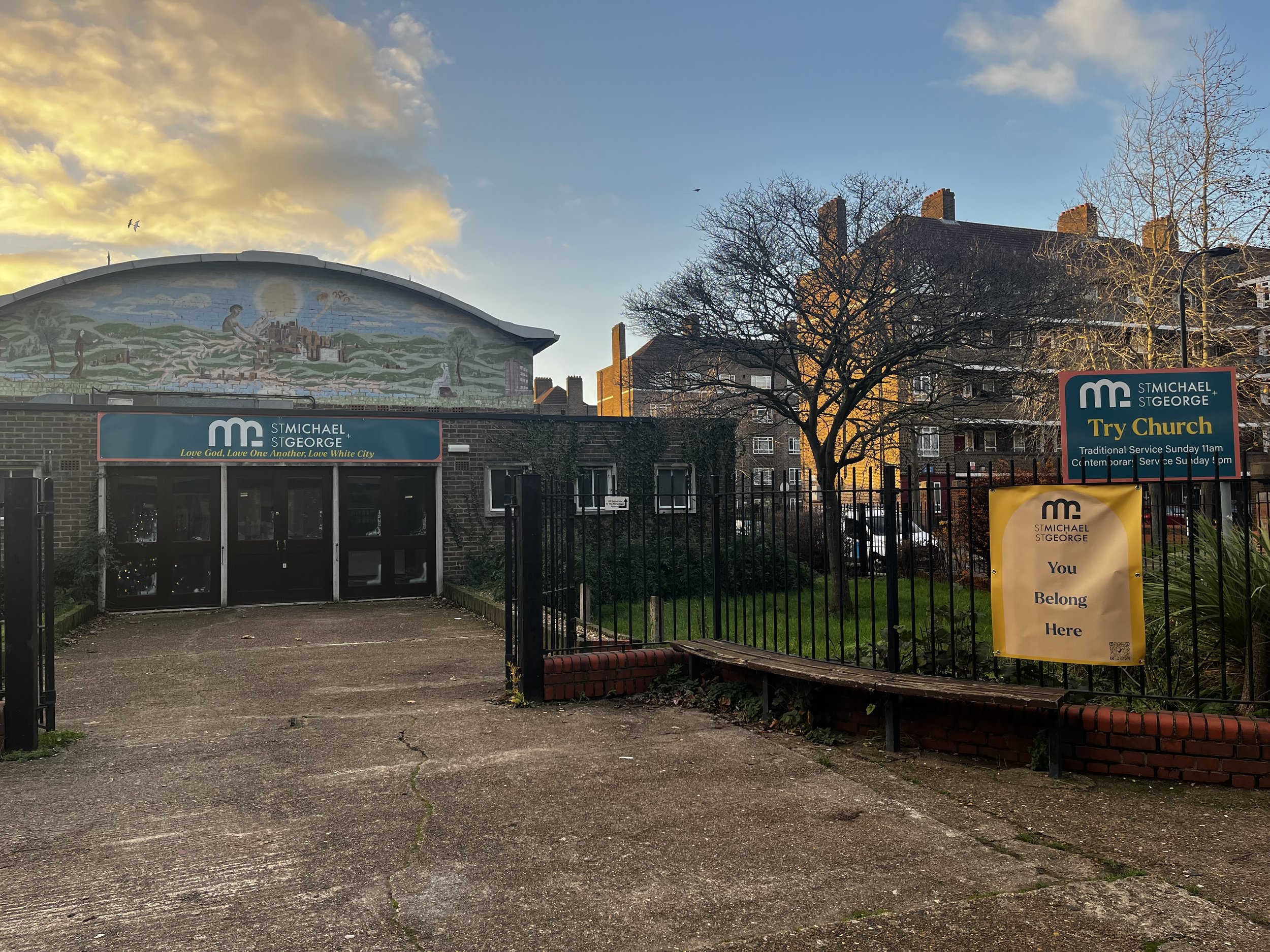 Exterior view of St. Michael & St. George church with signs indicating it is a church, and a small mural on the building, trees, parked cars, and residential buildings in the background.