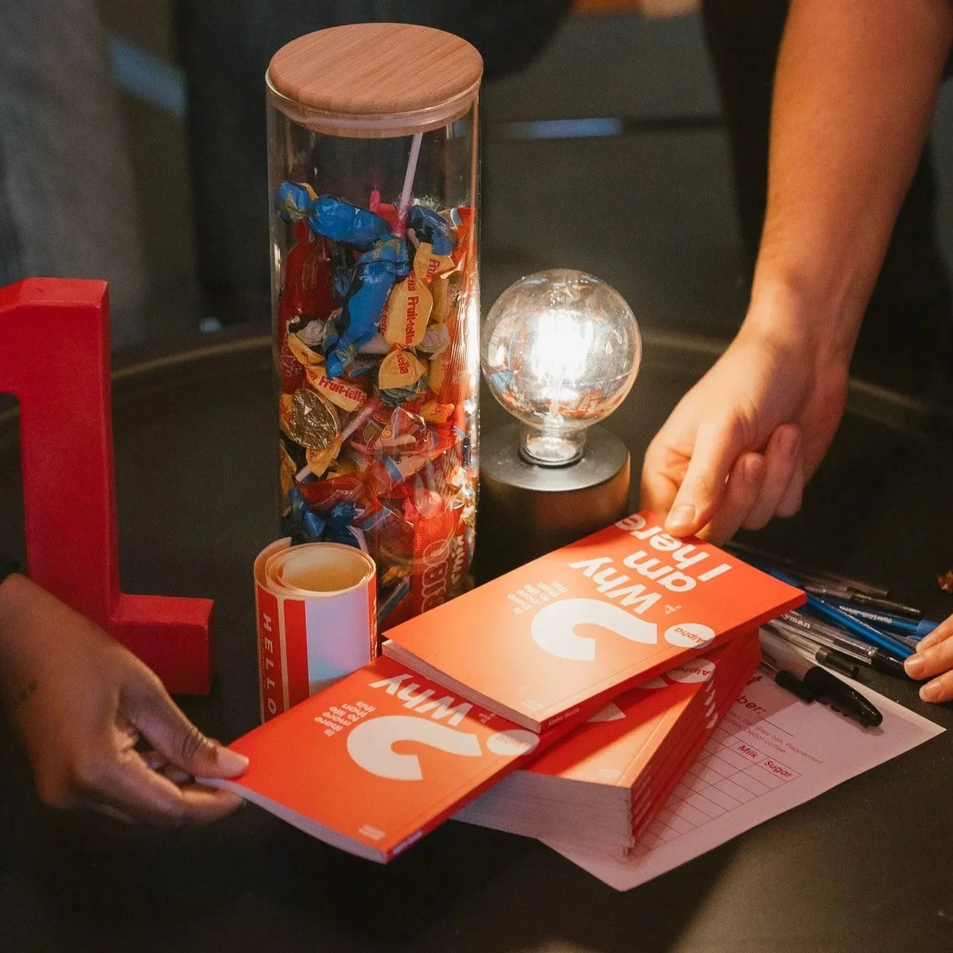 A person handling informational pamphlets labeled with a large white question mark, which read 'Why?'. The desk also features a jar filled with assorted wrapped candies, a small lighted decorative globe, a roll of stickers, and a sheet of paper with handwritten notes.