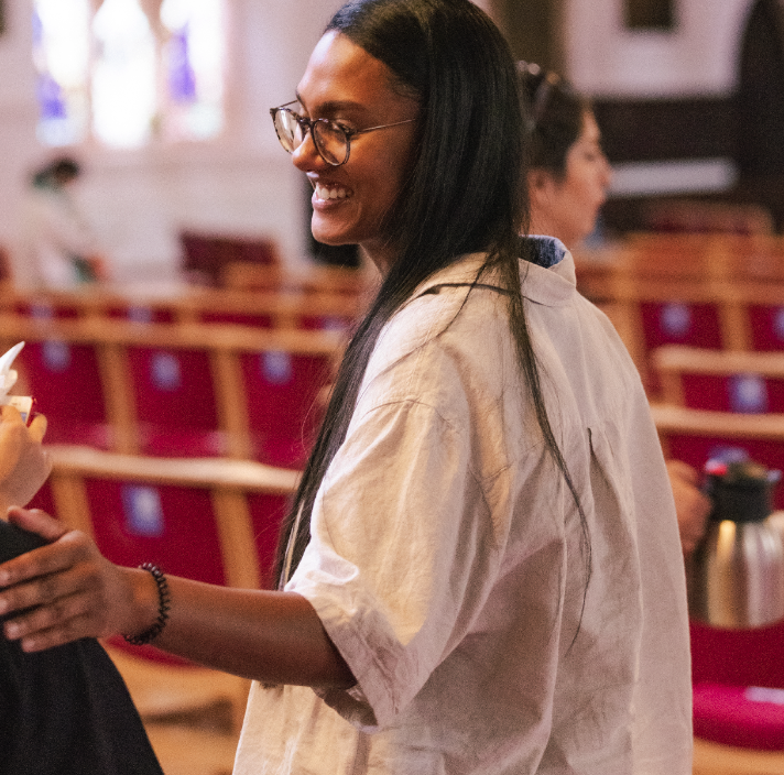 A young woman with long dark hair, wearing glasses and a light-colored shirt, smiling and holding a drink in a church or auditorium with red chairs in the background.
