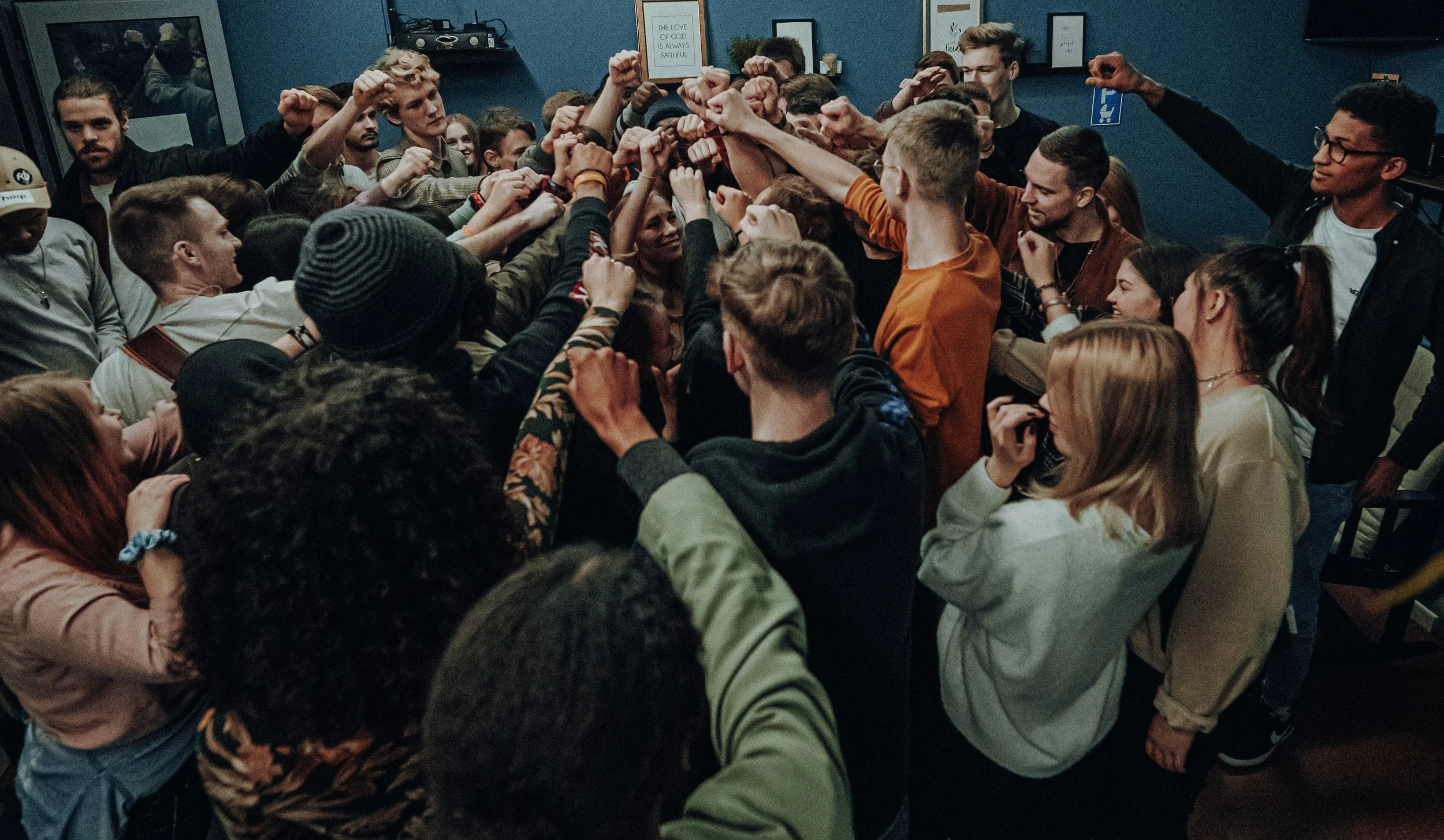 Group of young people forming a circle with their hands raised in the center, celebrating in a room with blue walls and framed pictures.