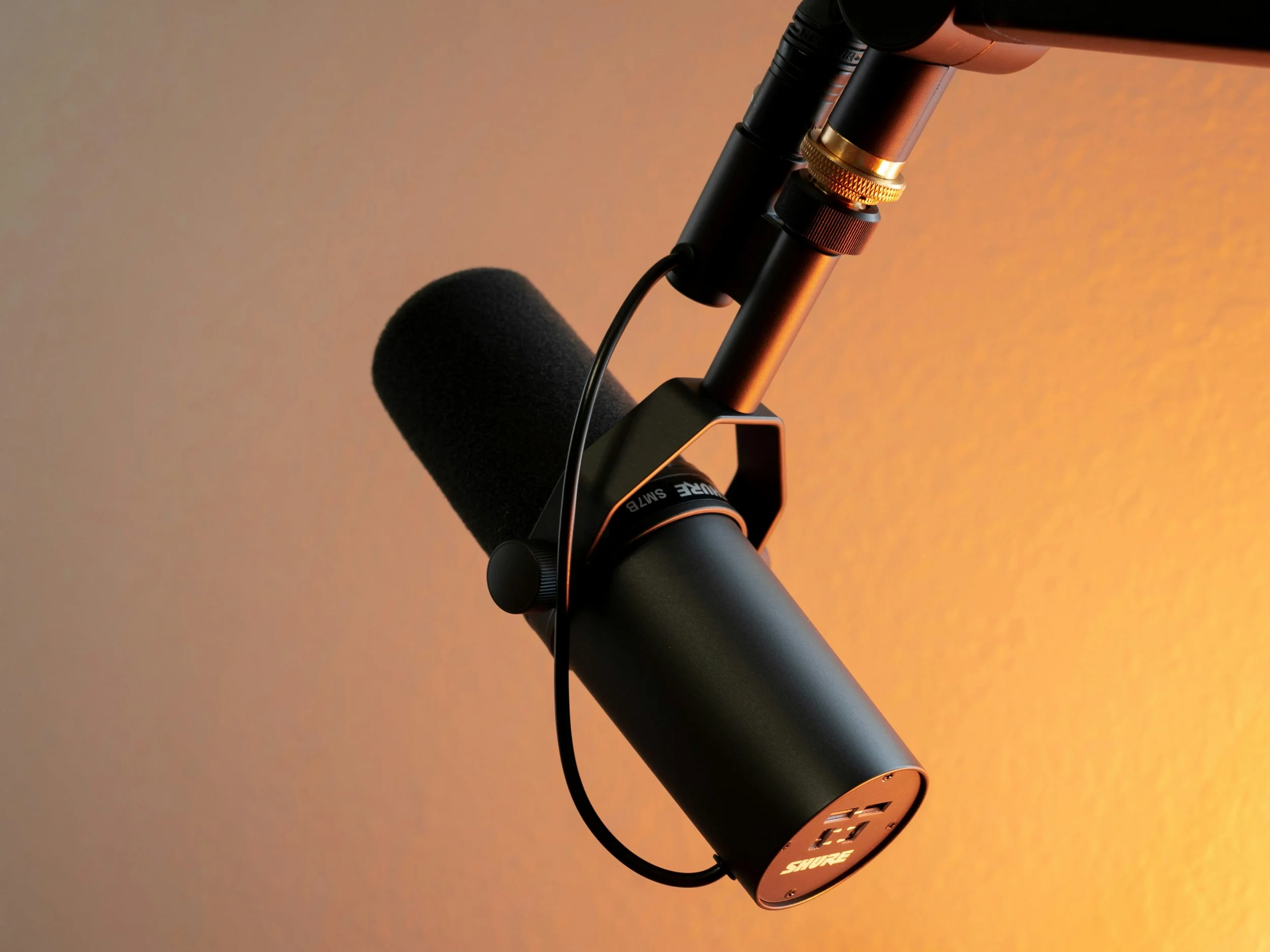Close-up of a professional microphone with black foam windscreen, mounted on a black adjustable arm against an orange gradient background.