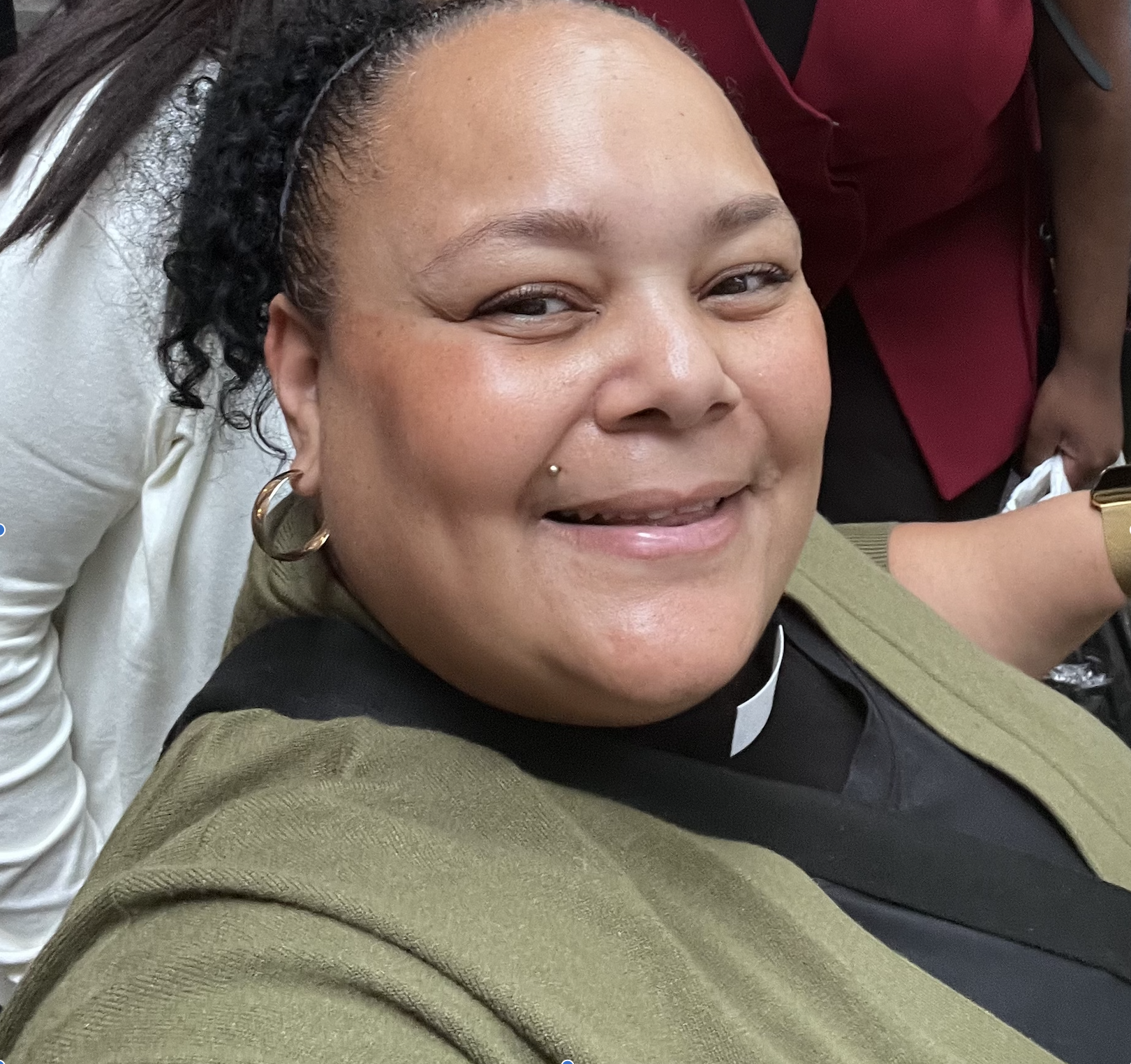 Close-up of a smiling woman with hoop earrings, a dermal pierce, wearing a clerical collar and olive green sweater, in a group setting.