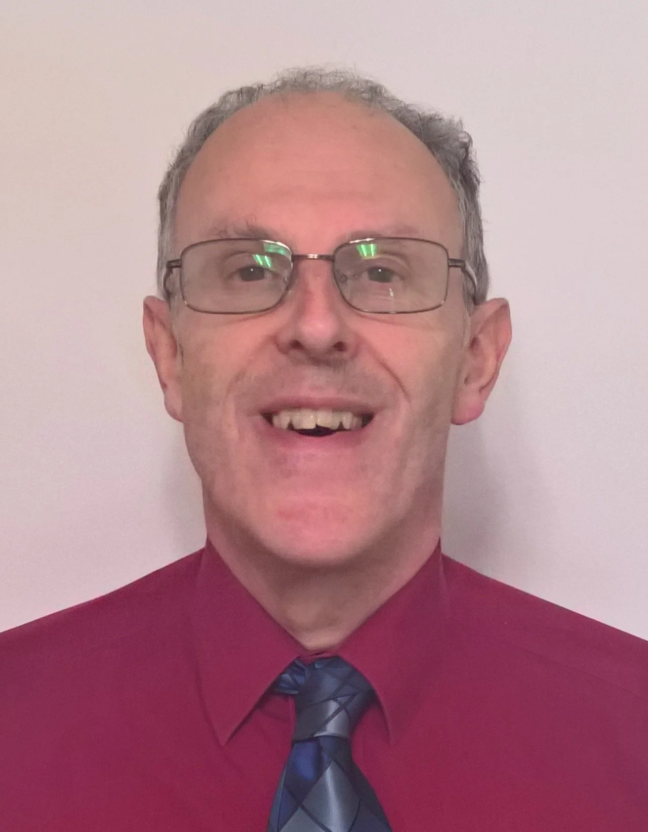 A middle-aged man with gray hair and glasses smiling in front of a plain light-colored wall, wearing a burgundy shirt and a patterned tie.