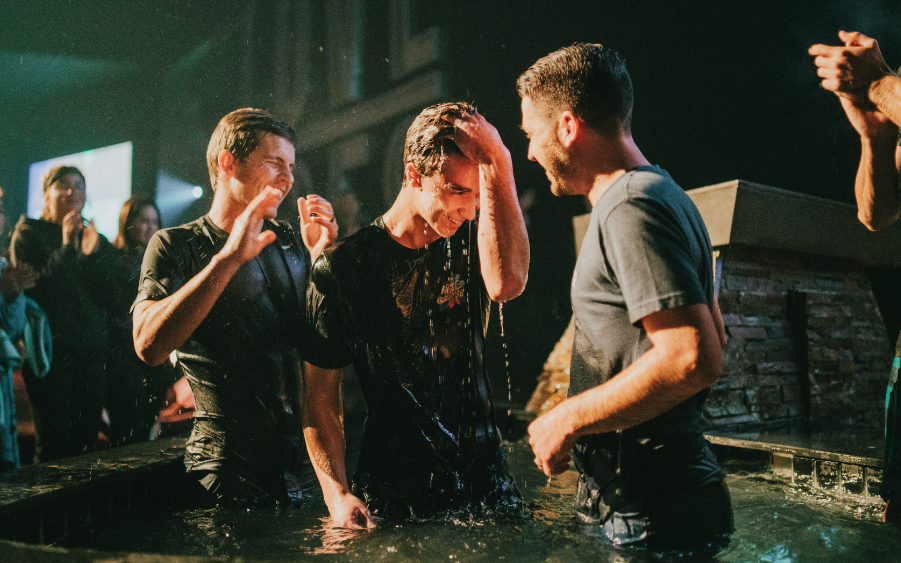 Three men, two of whom are smiling and one wiping water from his face, are standing in a baptismal pool during a baptism ceremony at a church or religious venue.