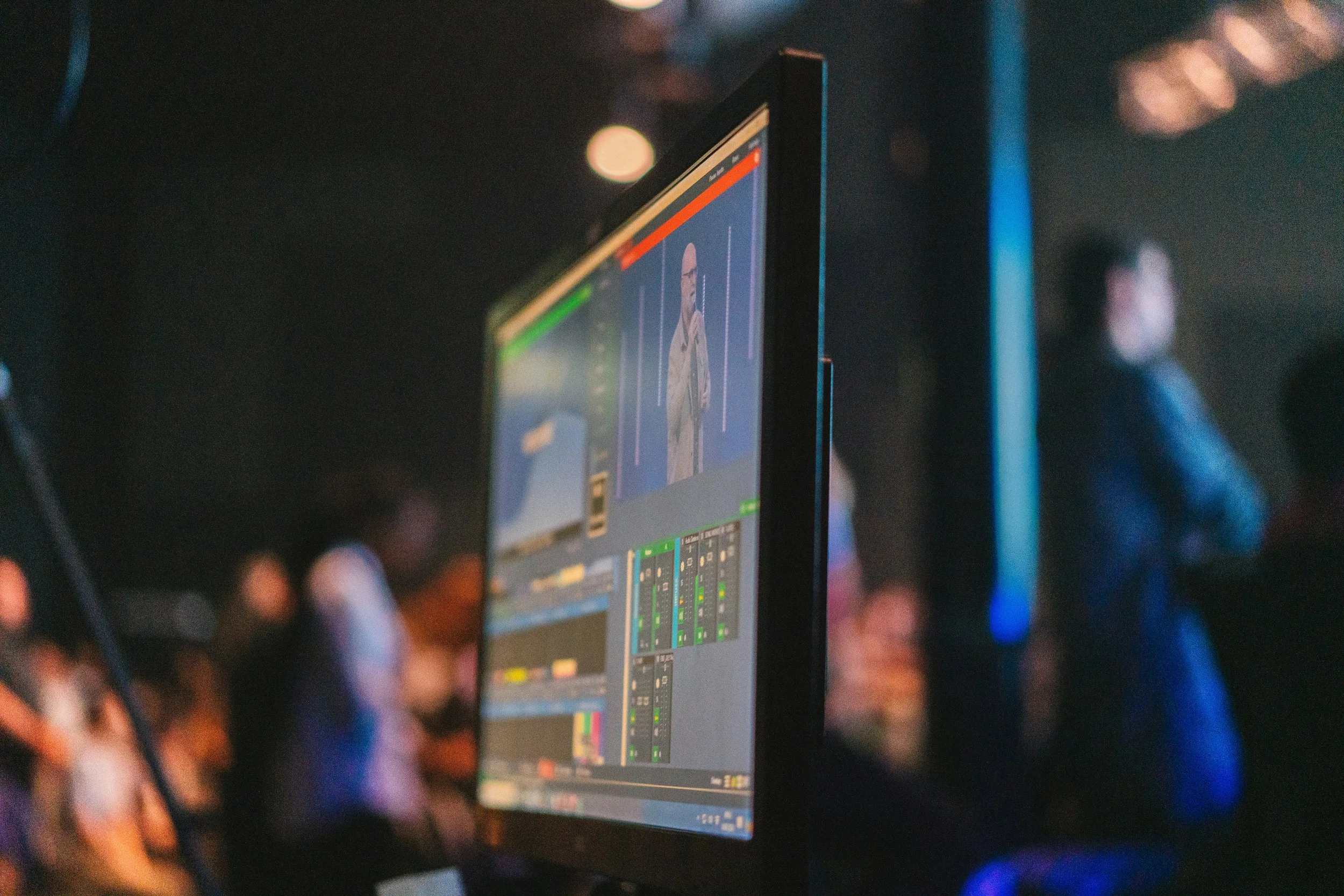 A computer monitor displaying a video production or streaming interface with a person on screen, in a dimly lit environment with blurred audience members in the background.