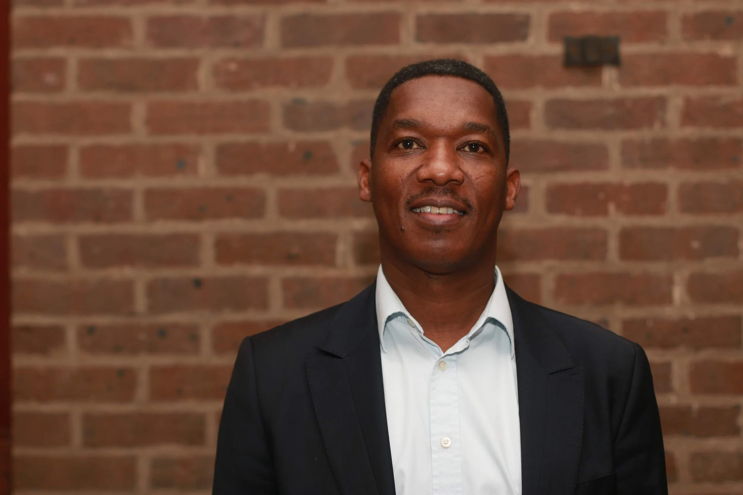 A man in a dark suit jacket and white dress shirt standing in front of a brick wall.