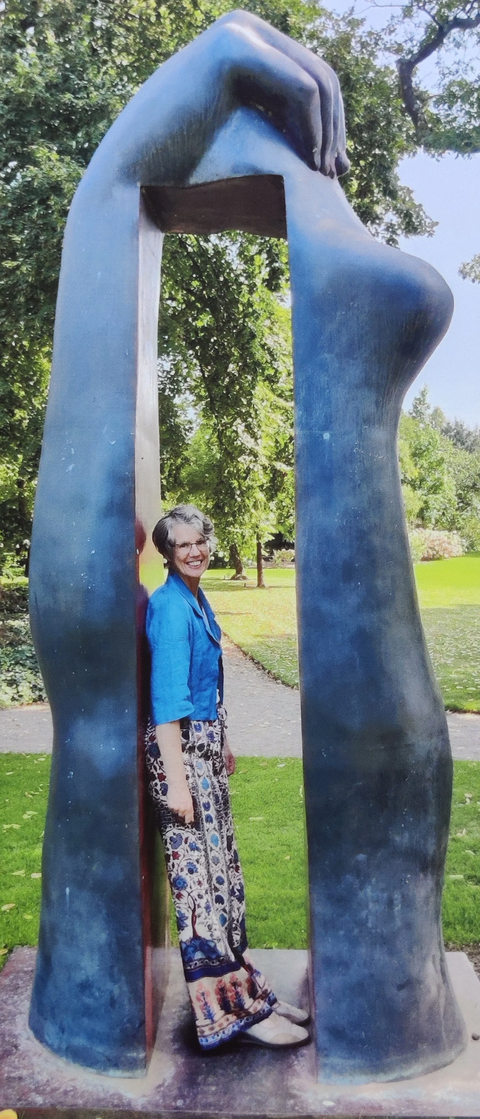 A woman with short gray hair, glasses, and a bright smile standing inside a large, abstract metal sculpture shaped like a head and hand, outdoors in a park with green trees and grass.
