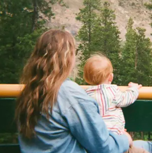 A woman holding a young child while looking at a landscape with trees and mountains.