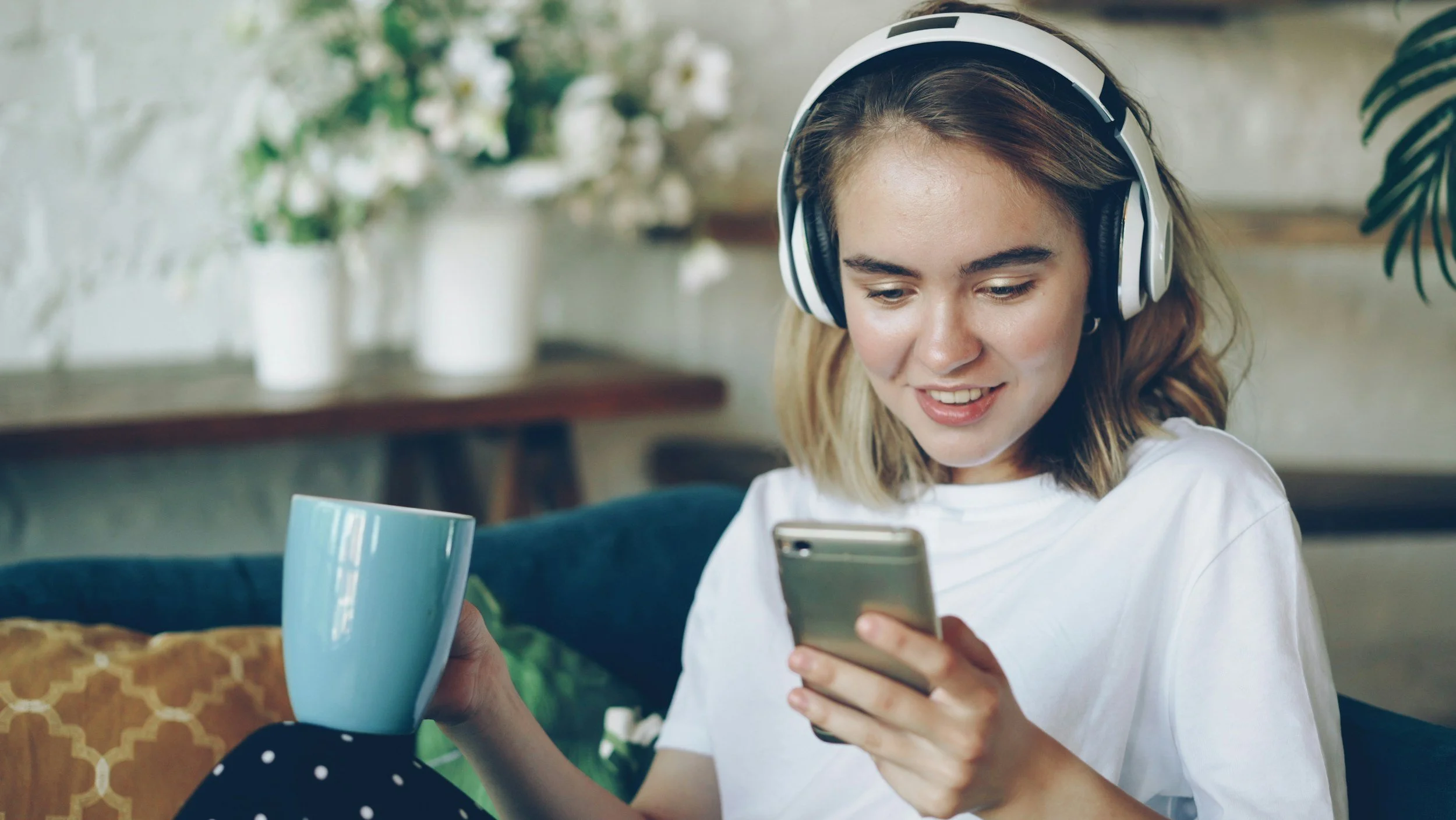 A young woman with headphones, smiling, holding a smartphone and a blue mug, sitting on a couch in a cozy room with plants in the background.