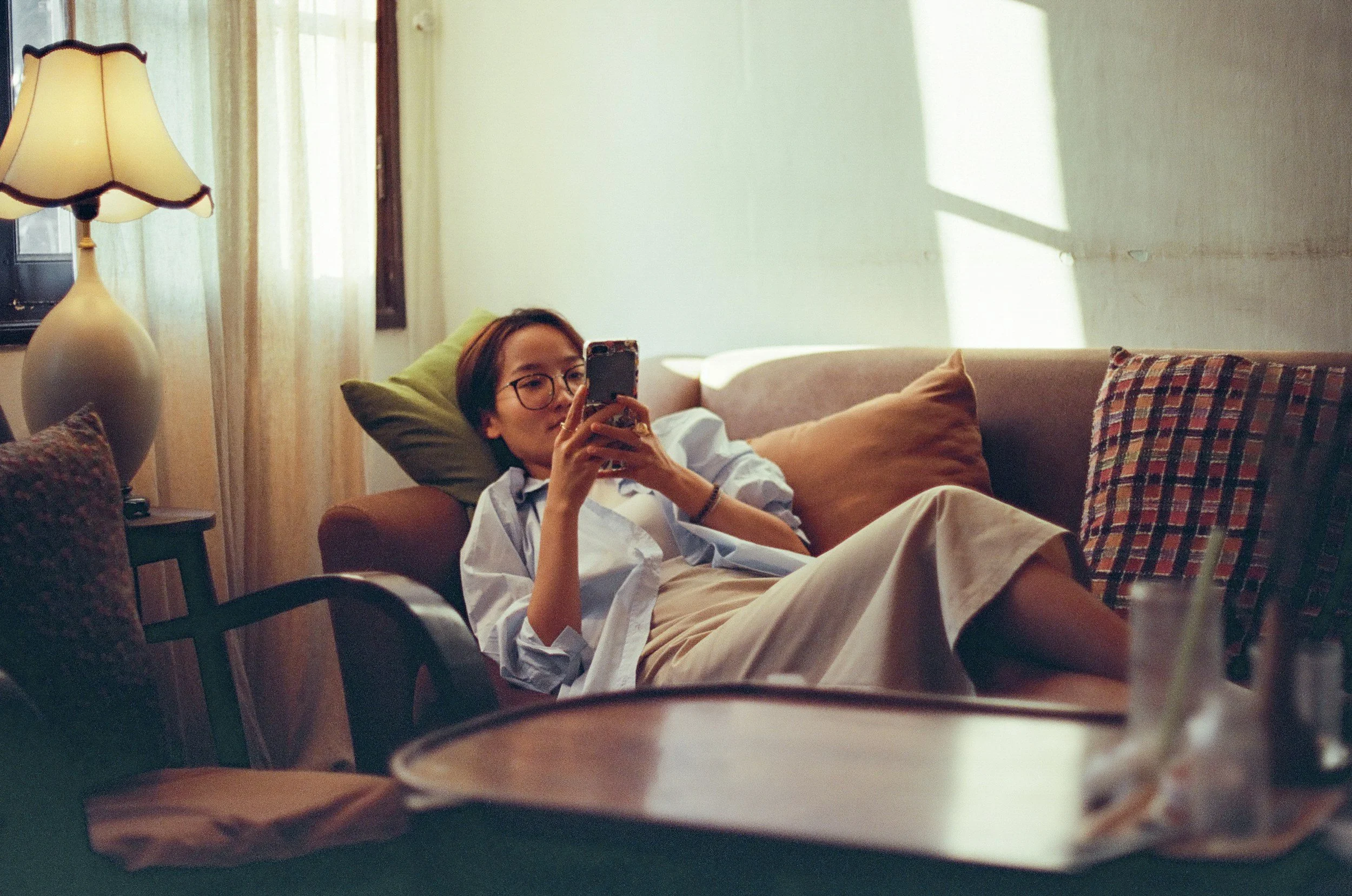 A woman with short brown hair and glasses is lying on a couch, looking at her phone. She is wearing a white shirt and beige pants. The living room has a lamp, a window with light-colored curtains, and several cushions on the sofa.