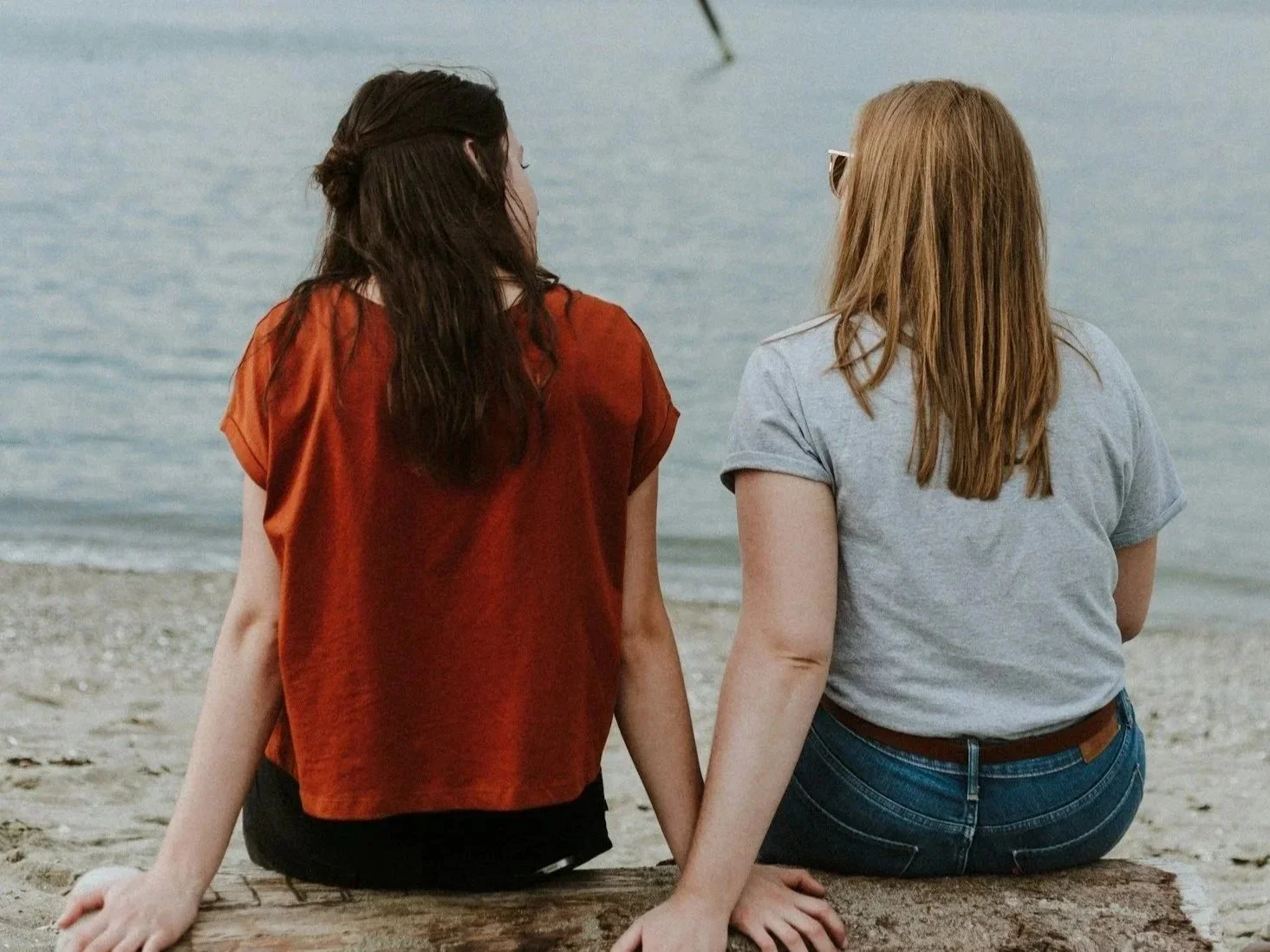 Two women from the Cove Collective infertility support community sitting side by side on a log at the beach, holding hands and facing the water, connecting on shared experience.