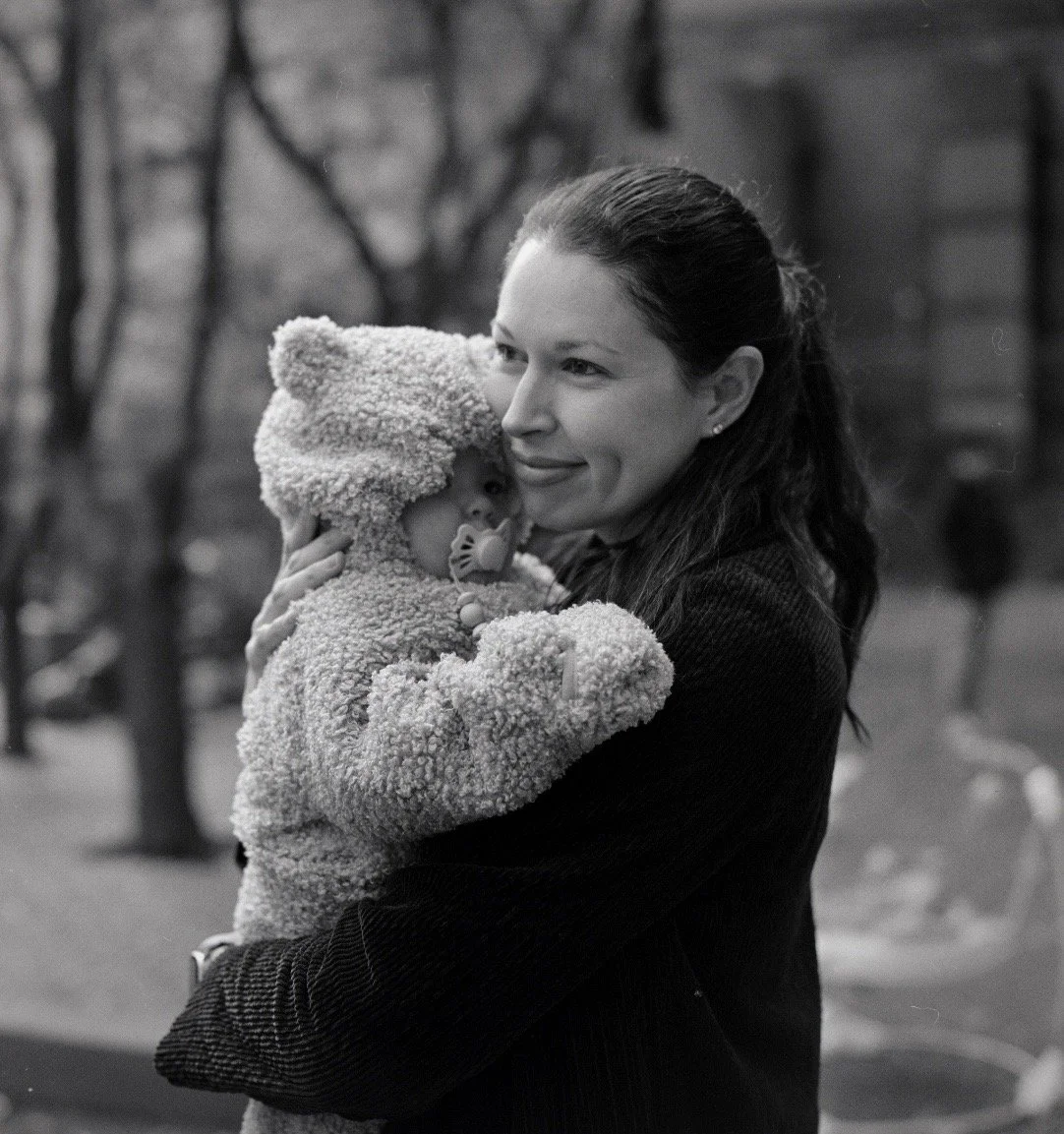 A woman holding a baby dressed in a teddy bear costume outdoors, with trees in the background.