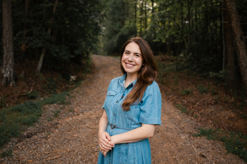 A woman with long brown hair, wearing a denim dress, standing on a forest trail and smiling at the camera.