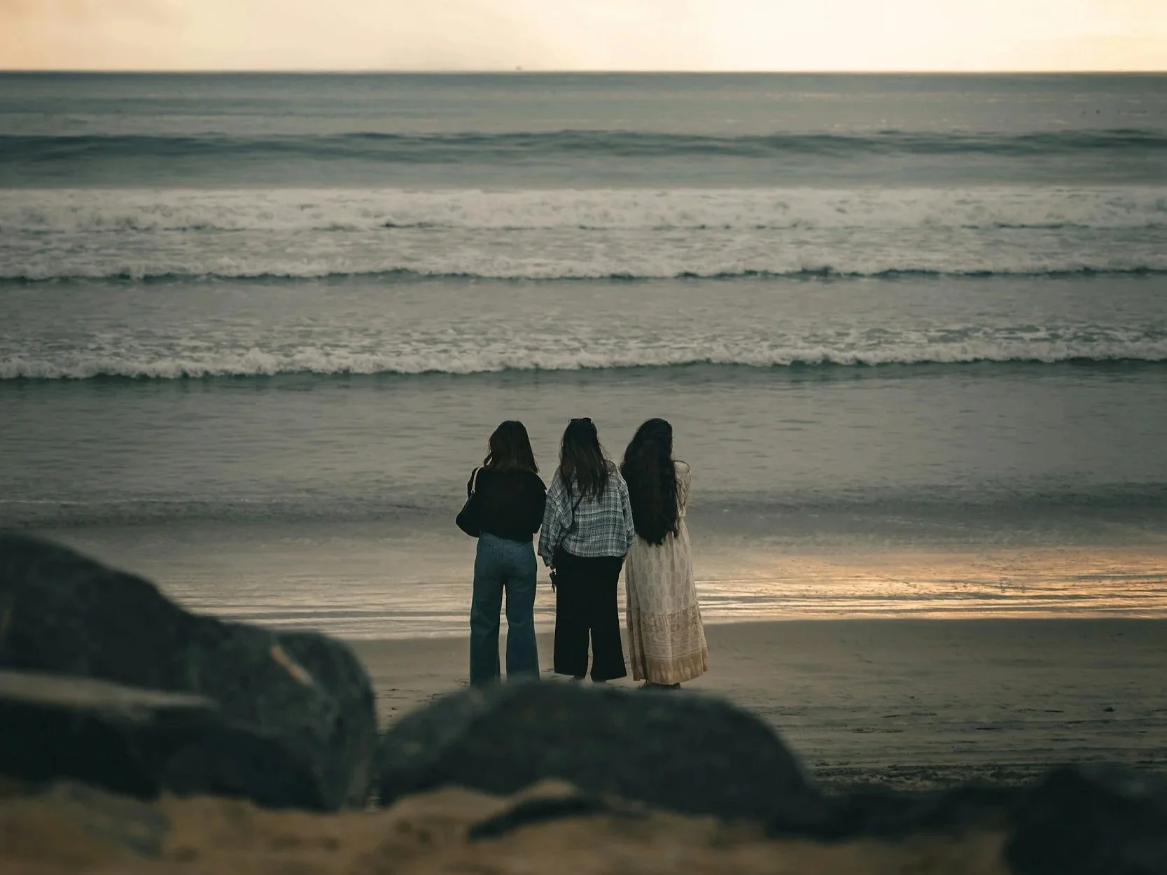 Three women standing on a beach at sunset looking at the ocean waves.