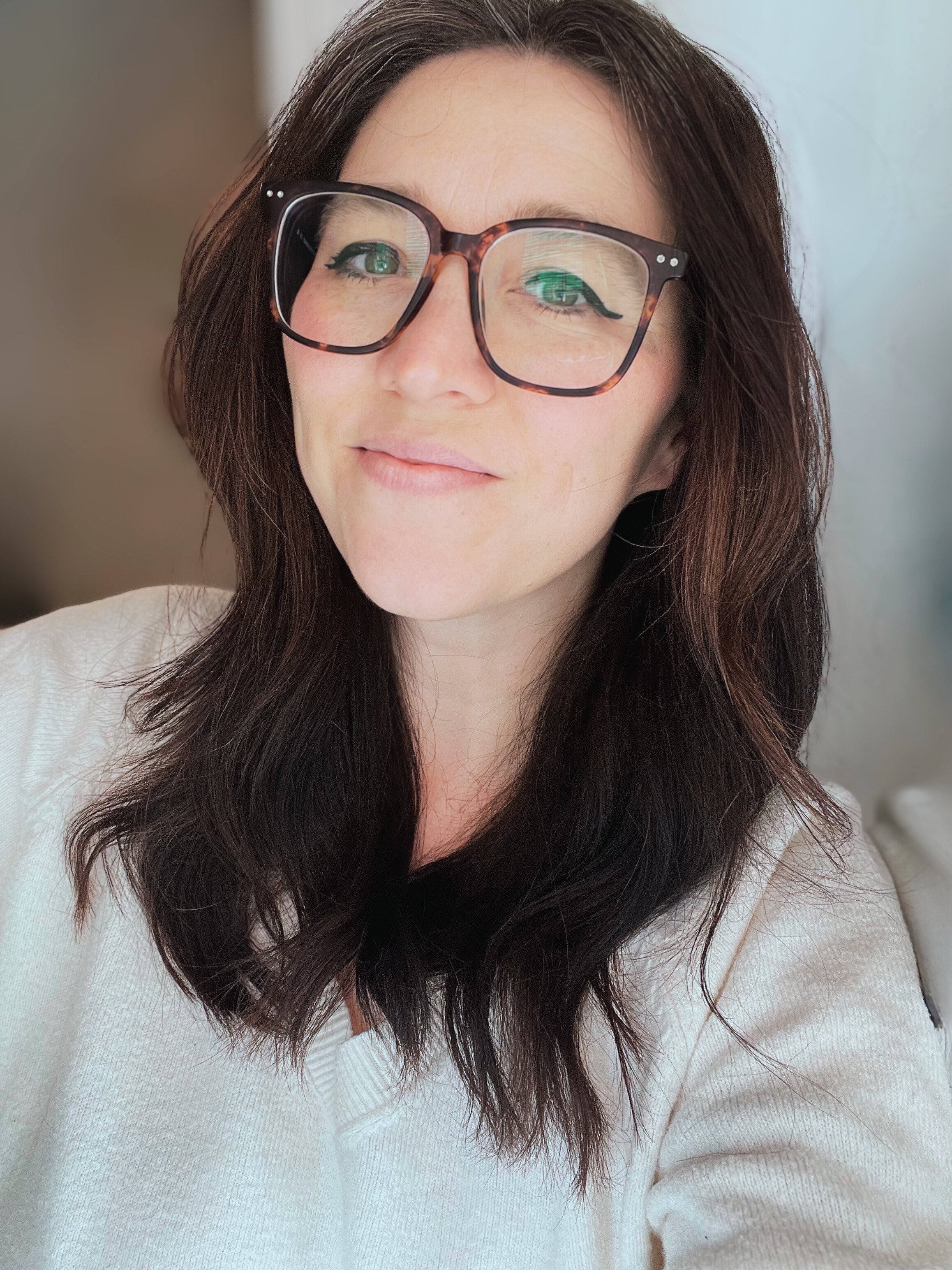 Close-up of a woman with dark brown wavy hair, wearing large tortoiseshell glasses, light makeup with eyeliner, and a light-colored top, smiling softly.