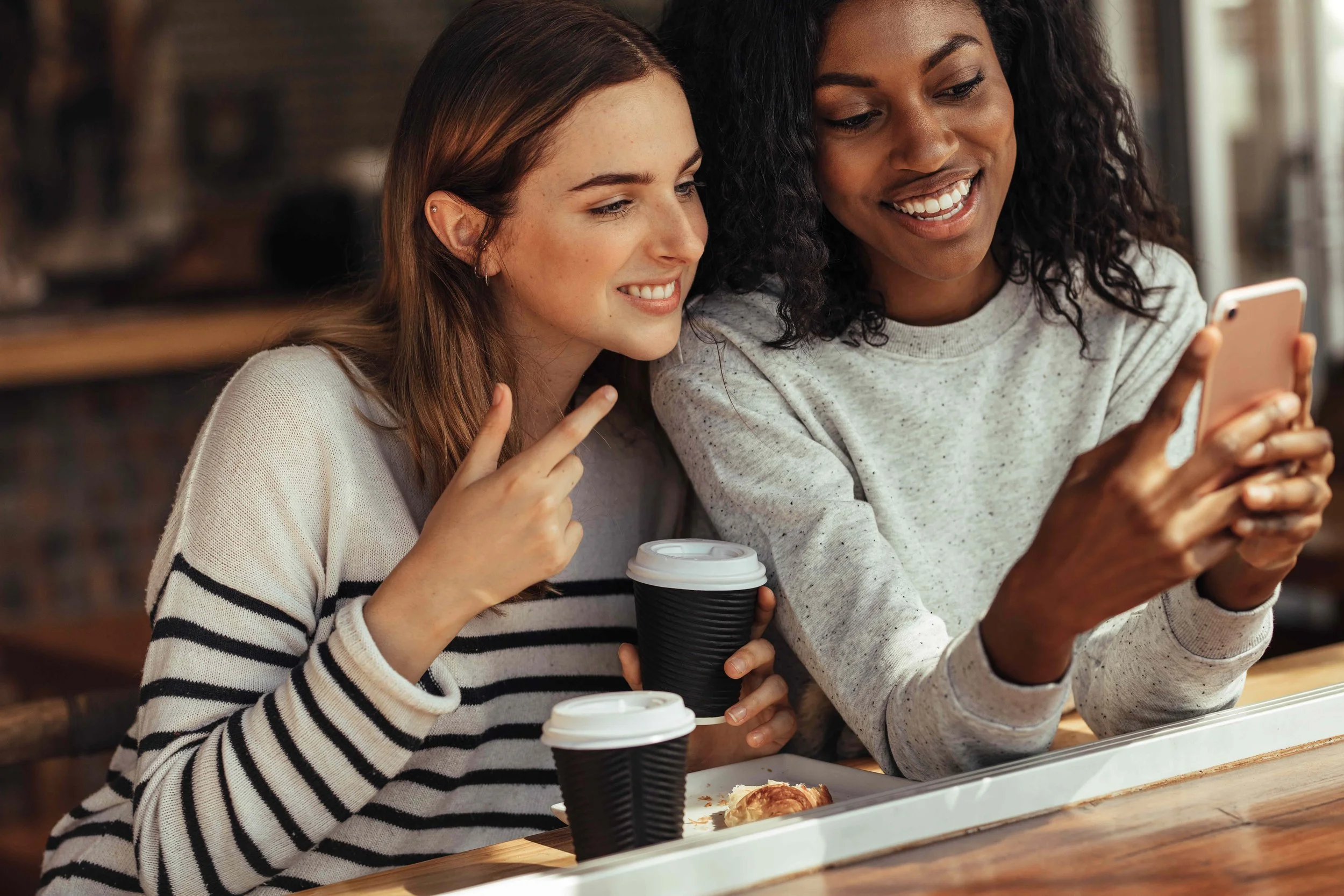 Two women sitting at a café countertop, looking at a smartphone and smiling, with coffee cups and a pastry on the table.