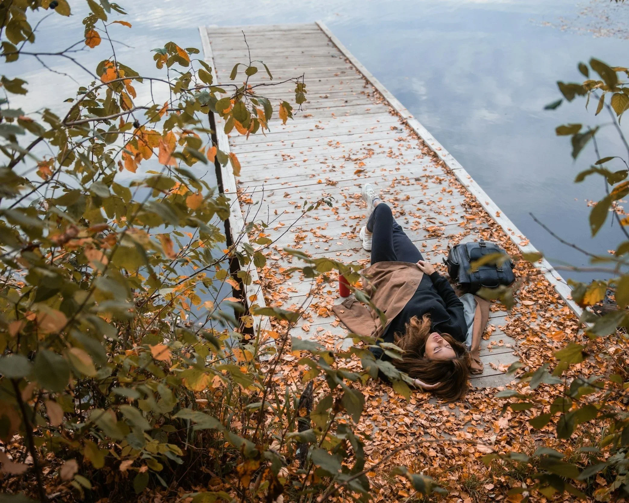 A woman lying on a wooden dock by a lake, surrounded by autumn leaves and foliage, with her head resting on her hand and looking at the camera.