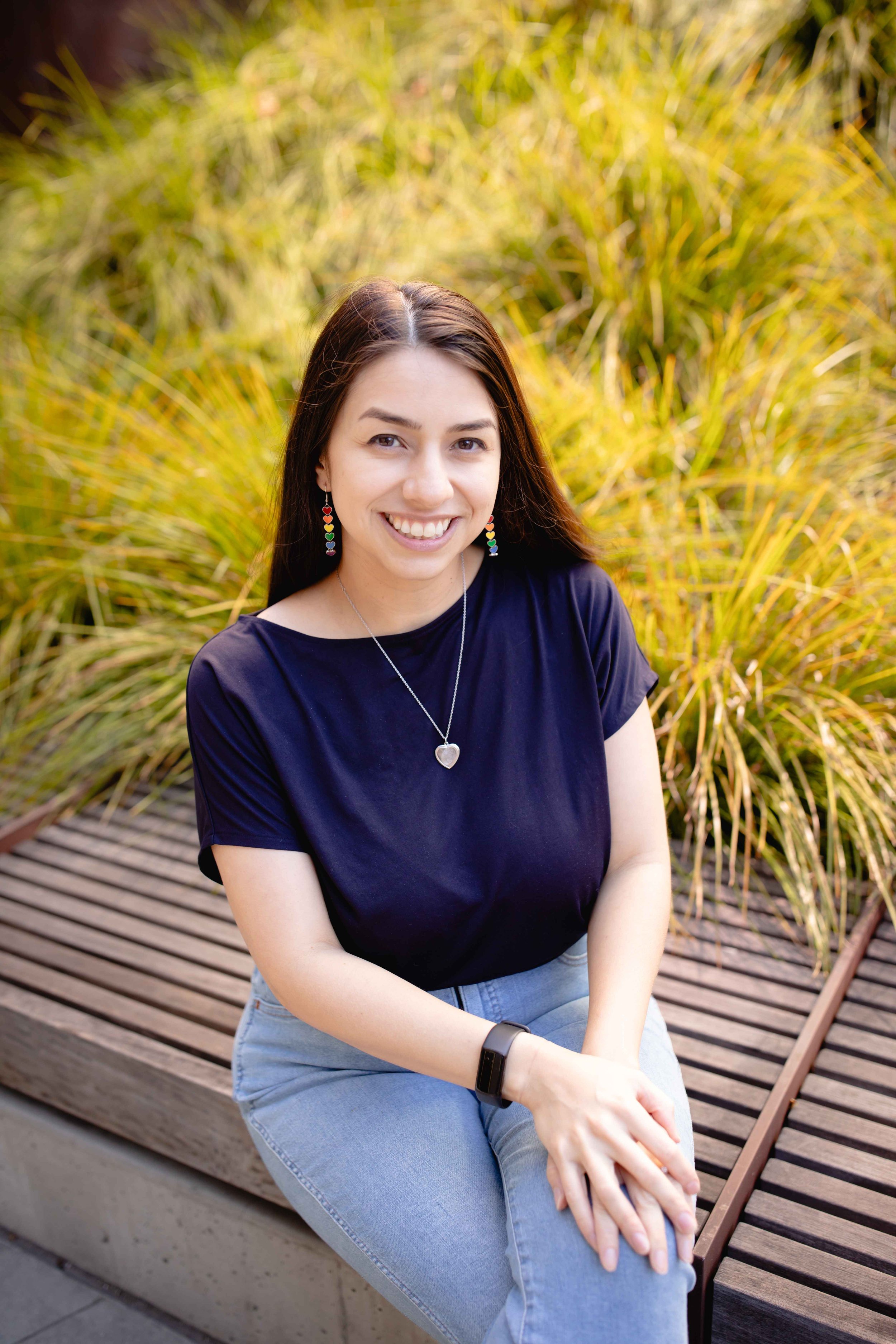 A woman with long dark hair smiling and sitting on a wooden bench outdoors, wearing a black t-shirt, blue jeans, colorful earrings, a silver heart necklace, and a black smartwatch, with yellowish tall grass in the background.