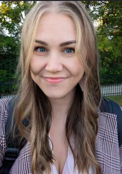 Young woman with blonde hair and blue eyes smiling outdoors with trees in the background.