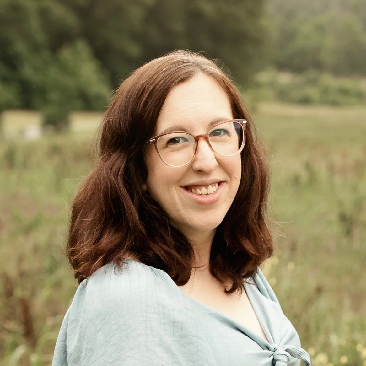 A woman with shoulder-length brown hair, glasses, and a light blue blouse smiling outdoors with a blurred natural background of trees and water.