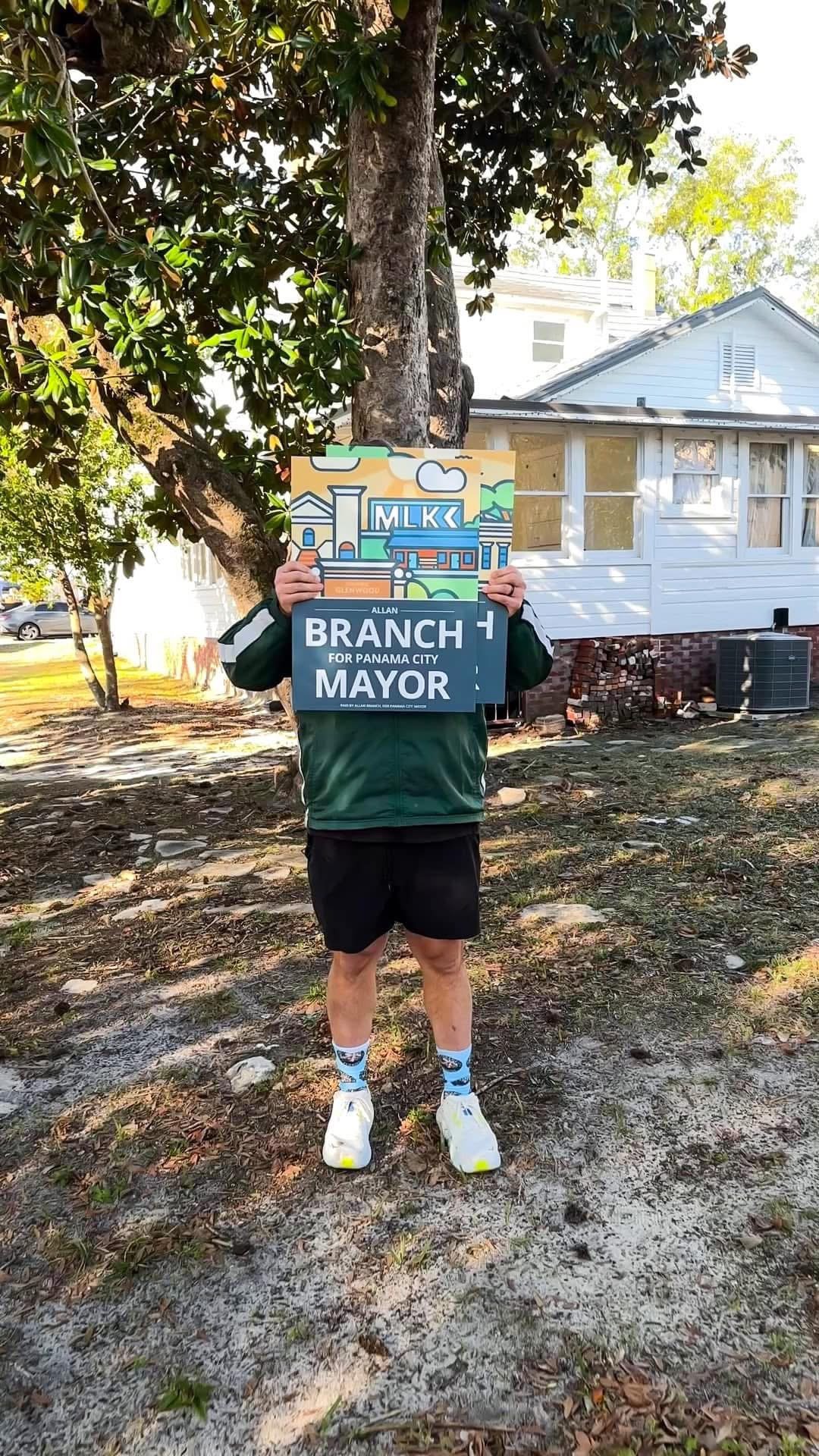 Person standing outdoors holding a political campaign sign blocking their face. The sign promotes Branch for Panama City mayor. The person is wearing a green jacket, black shorts, white running shoes, and white Socks with a design. Background includes a tree, house, and parked cars.