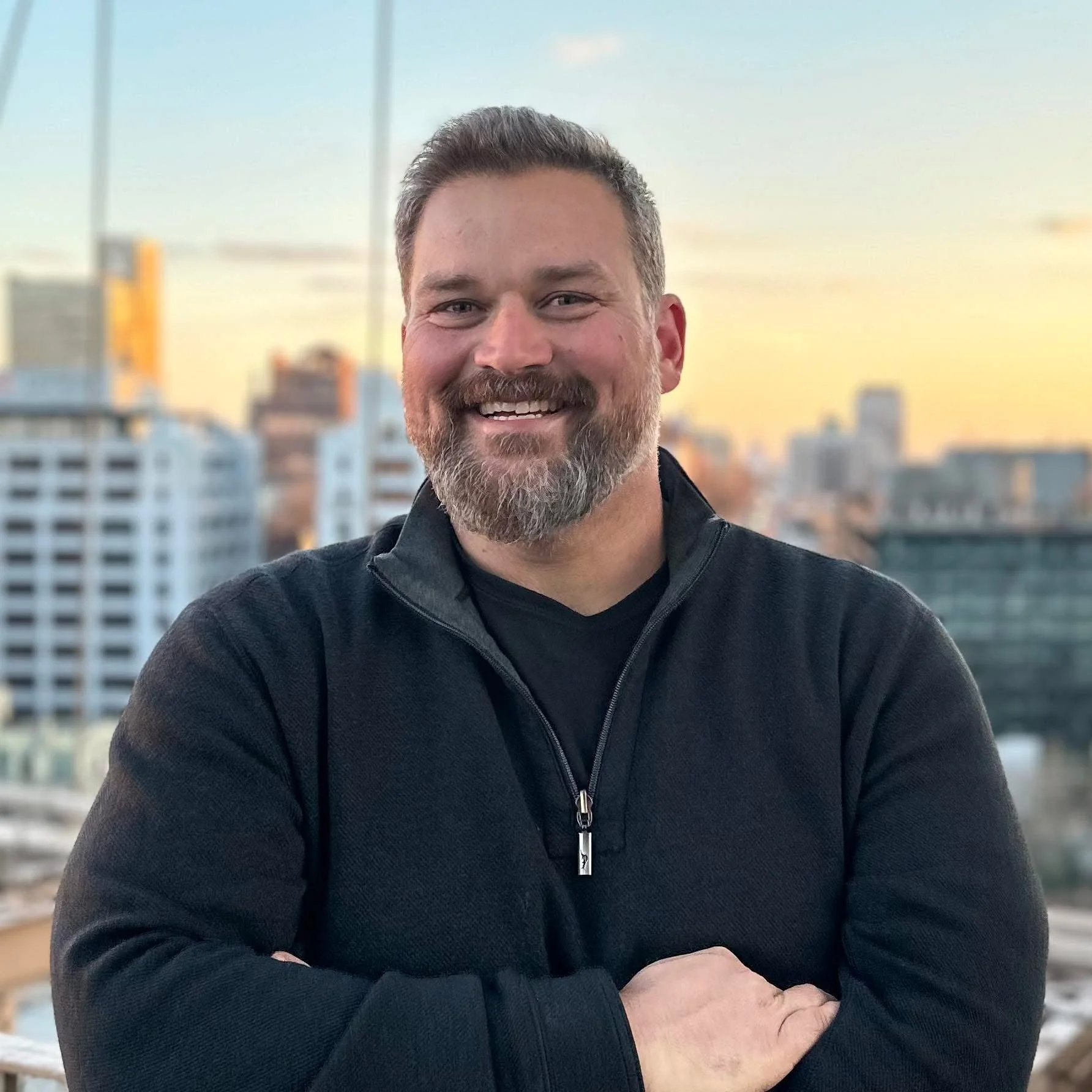 A smiling man with a beard and short hair, wearing a black zip-up jacket, standing with arms crossed against a city skyline at sunset.