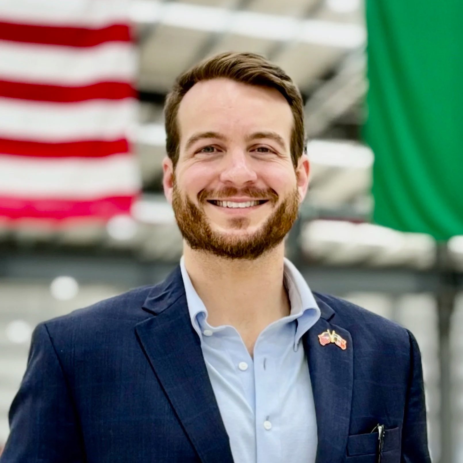 A smiling man with brown hair and beard, wearing a blue blazer and a light blue shirt, standing in front of American and green flags.