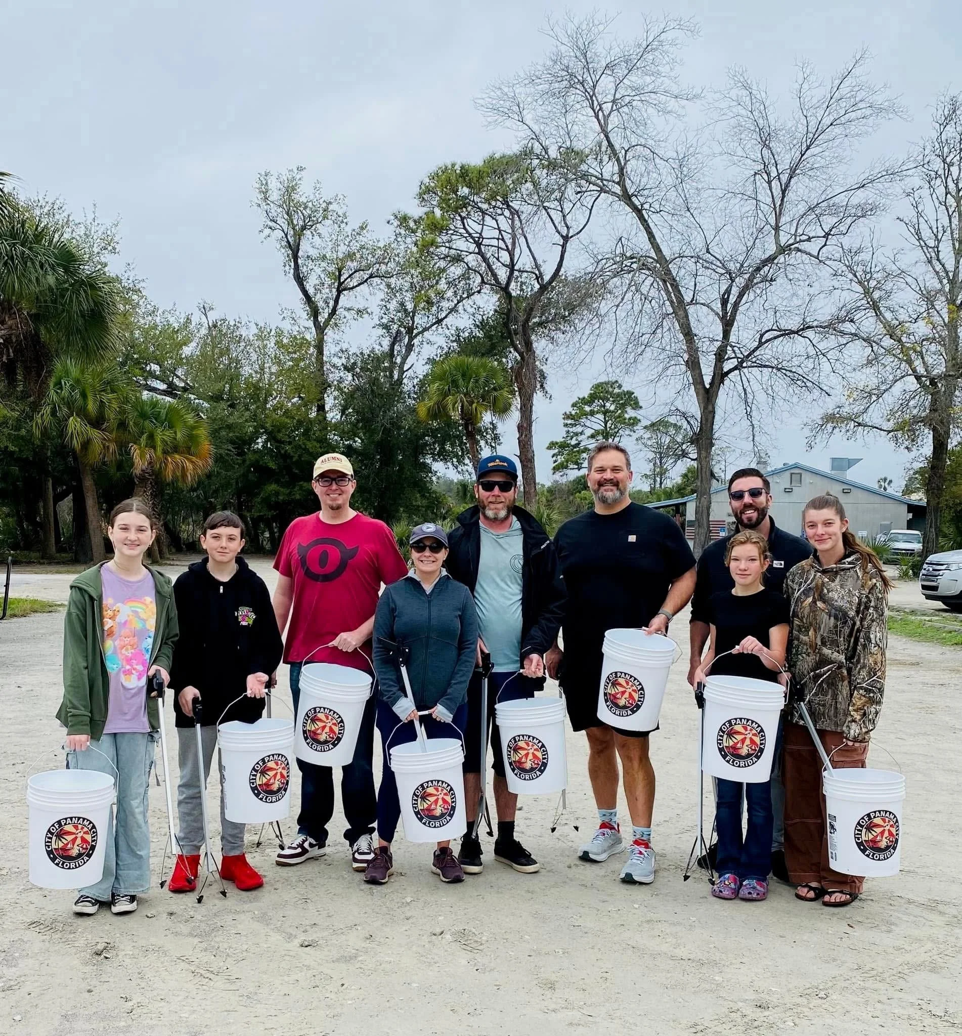 Group of nine people standing on a dirt trail outdoors, each holding a white bucket with a city of Panama logo, surrounded by trees and overcast sky.