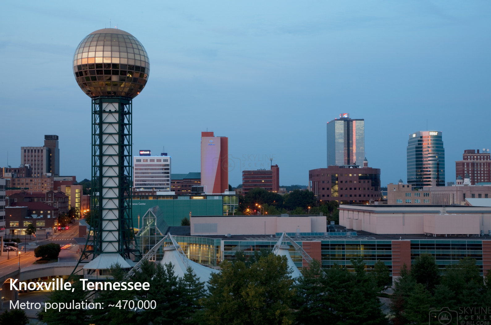 Dusk view of downtown Knoxville, Tennessee, featuring a spherical observation tower, several modern high-rise buildings, and green trees in the foreground.