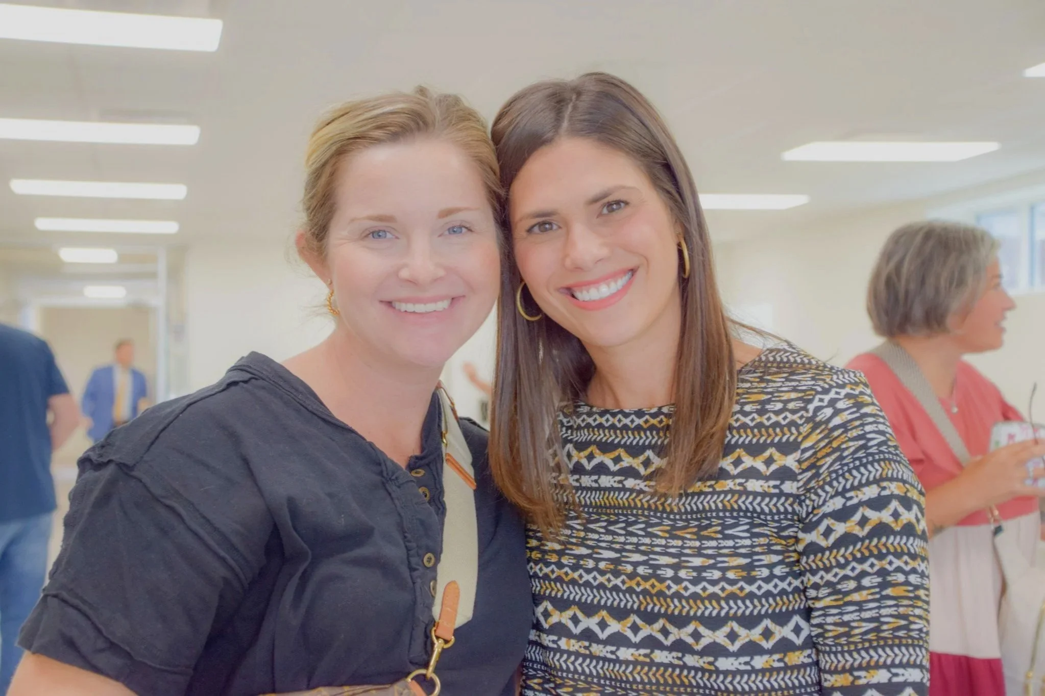 Two women smiling and posing together at an indoor event, with other people in the background.