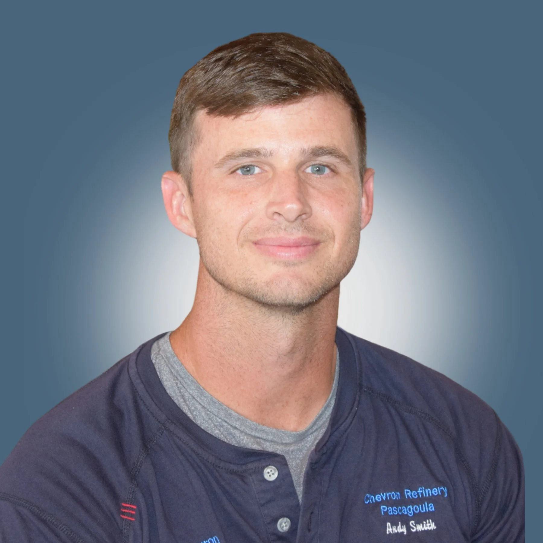 A portrait of a young man with short brown hair and blue eyes smiling, wearing a navy blue shirt with embroidery reading "Chevron Refinery Pascagoula Andy Smith" against a gray gradient background.