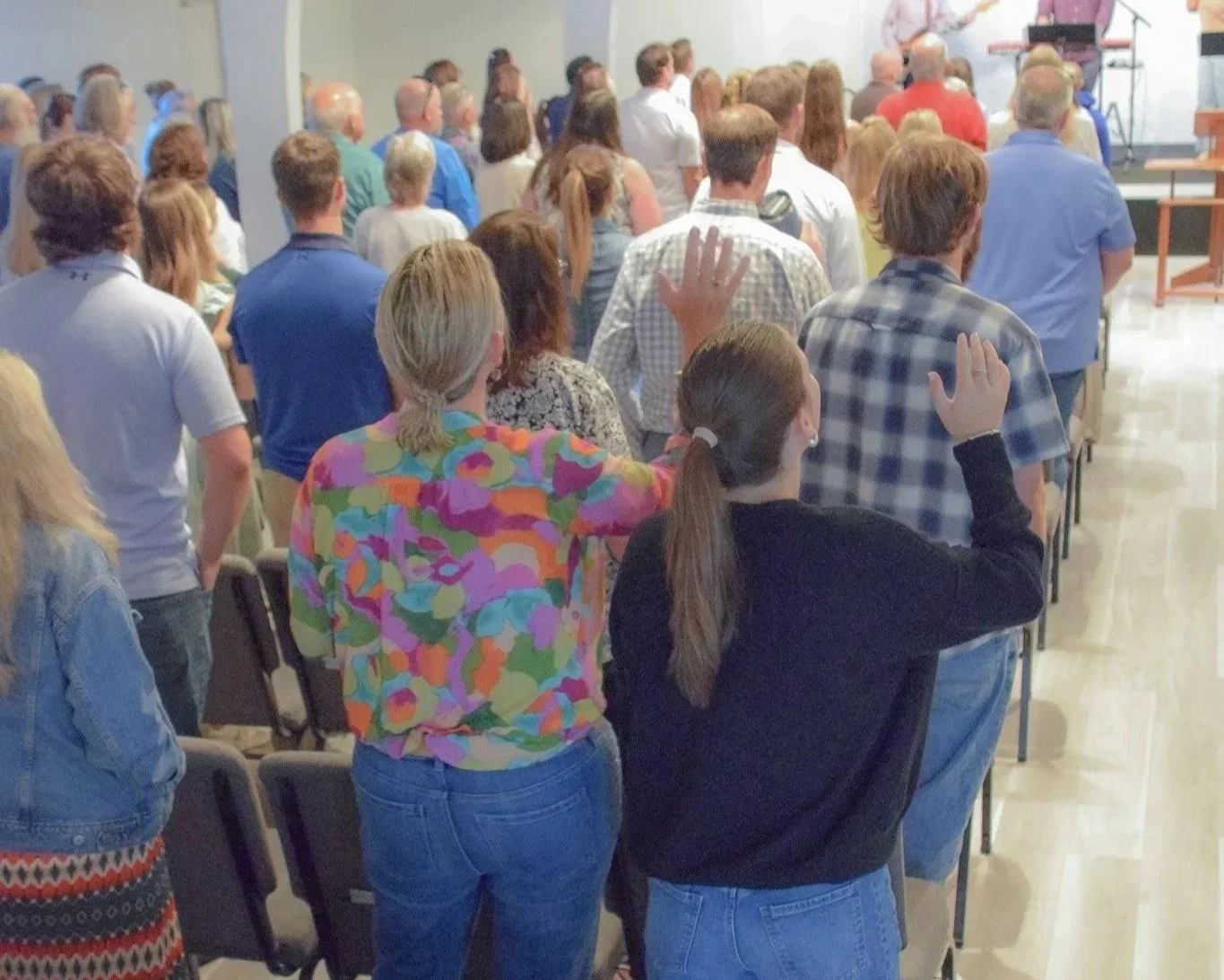 A large group of people in a religious or community gathering, standing with many raising their right hands during a ceremony or prayer session in an indoor hall.