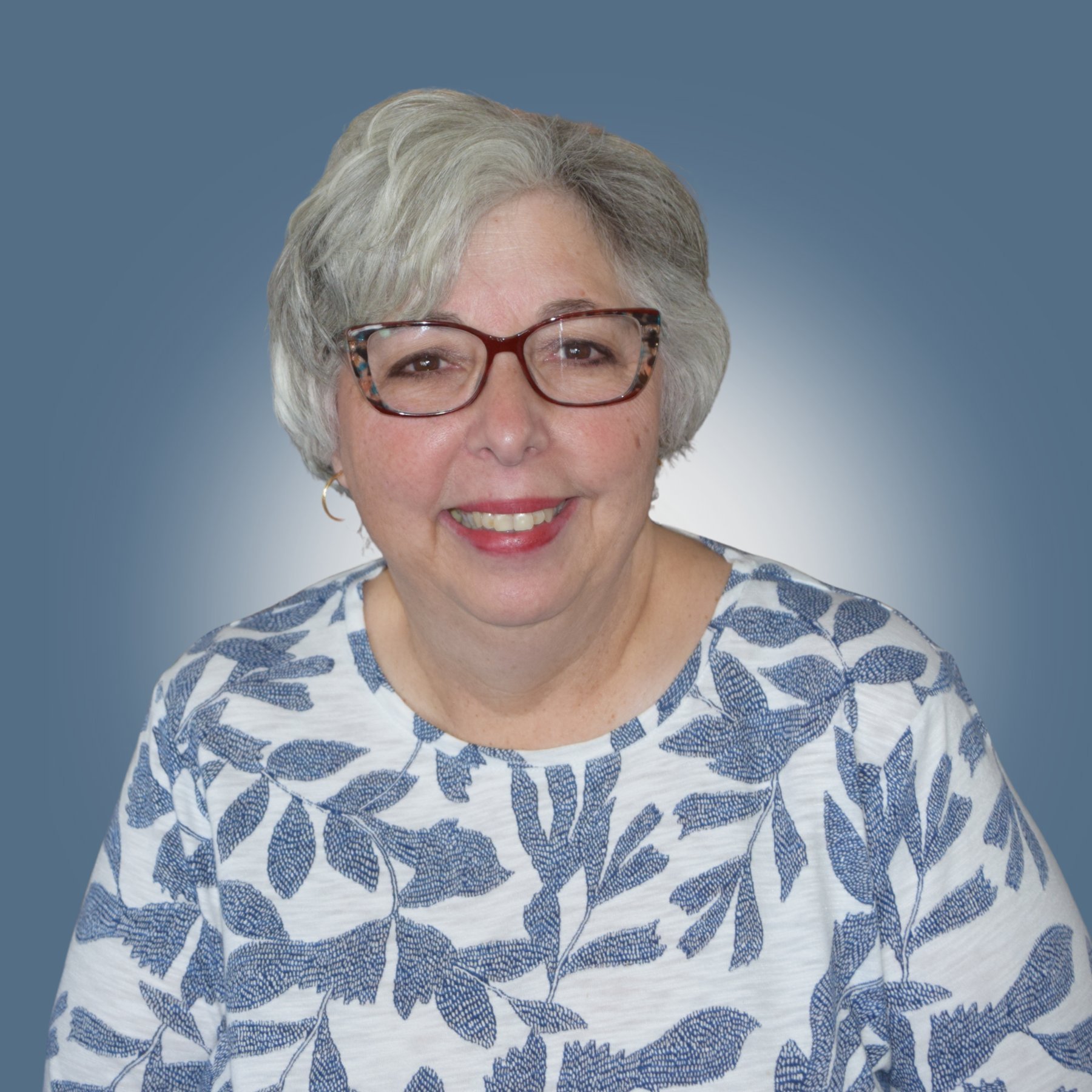 Smiling elderly woman with glasses and short gray hair, wearing a white blouse with blue leaf patterns, posed against a blue gradient background.