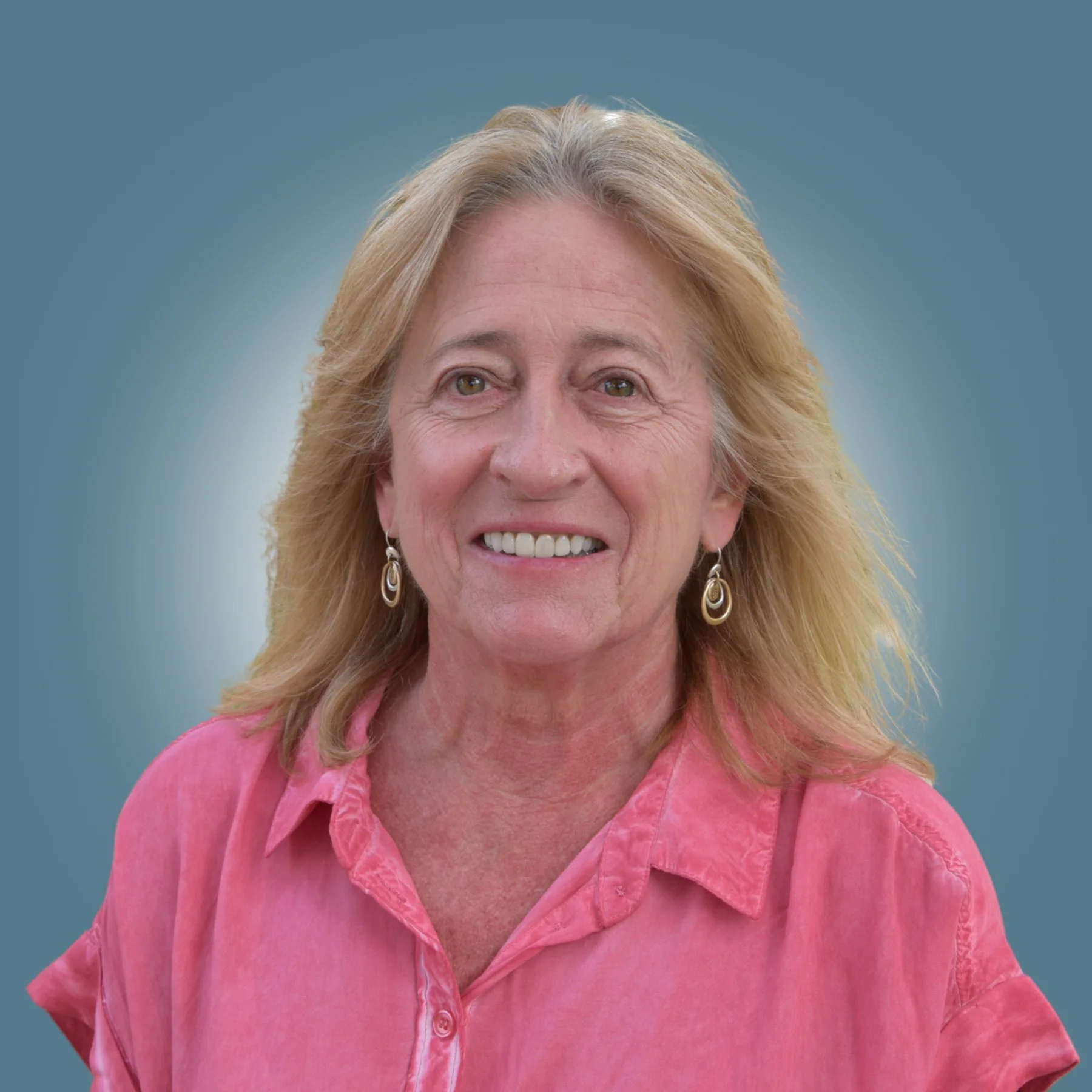 A smiling middle-aged woman with blonde hair, wearing a pink blouse and earrings, against a plain teal background.