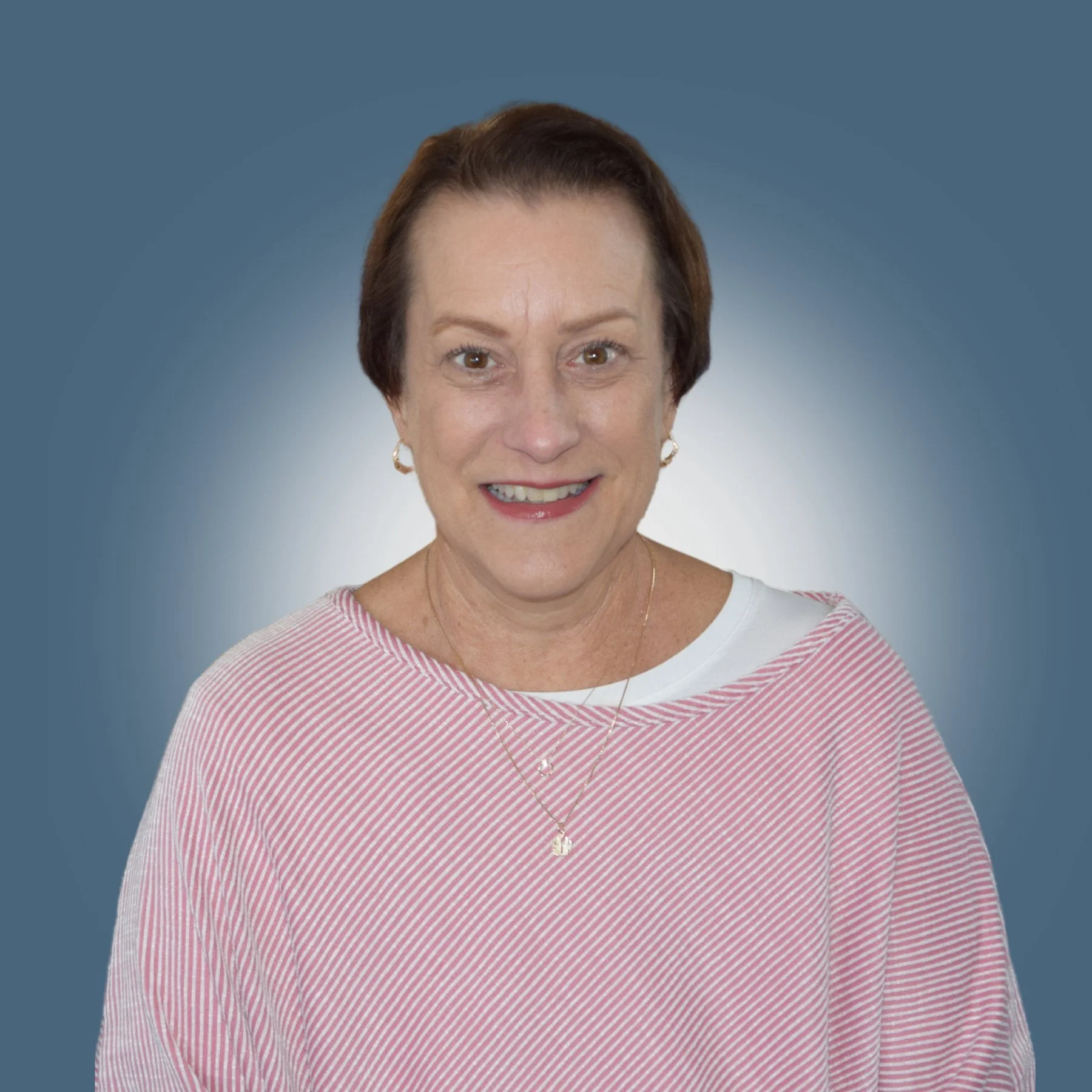 Portrait of an older woman with short brown hair, smiling, wearing a pink and white striped top with hoop earrings, a delicate necklace, and a white shirt underneath, against a gradient blue background.
