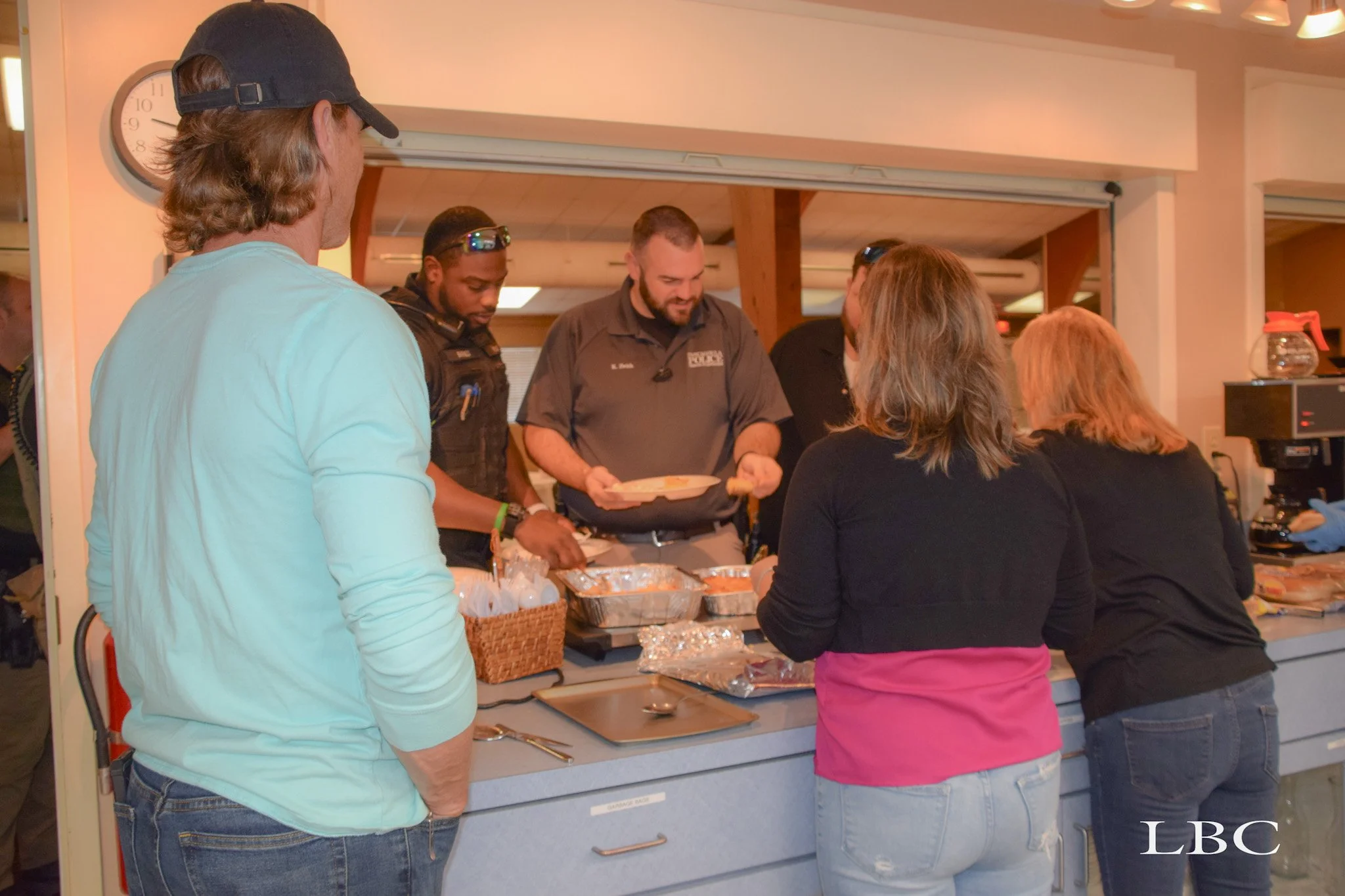 People standing in line at a buffet-style food counter, being served by staff members. The staff is behind the counter with trays of food, and the customers are on the other side, waiting to be served.