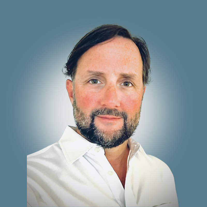 Portrait of a middle-aged man with dark hair and a beard, wearing a white collared shirt, against a plain background.