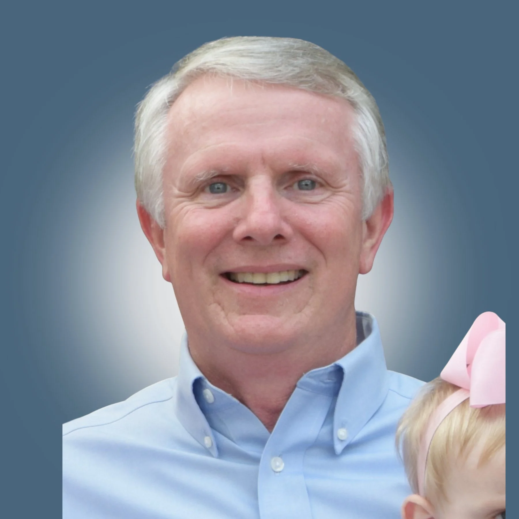 A middle-aged man with silver hair wearing a light blue button-up shirt, smiling with a slight gap in his teeth, against a blurred blue background.