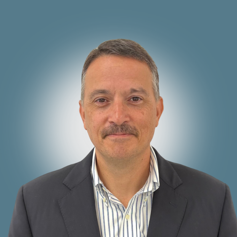 A man with short brown hair, a mustache, and a slight smile wearing a dark suit jacket over a striped dress shirt, standing against a blue background with a soft white glow behind his head.