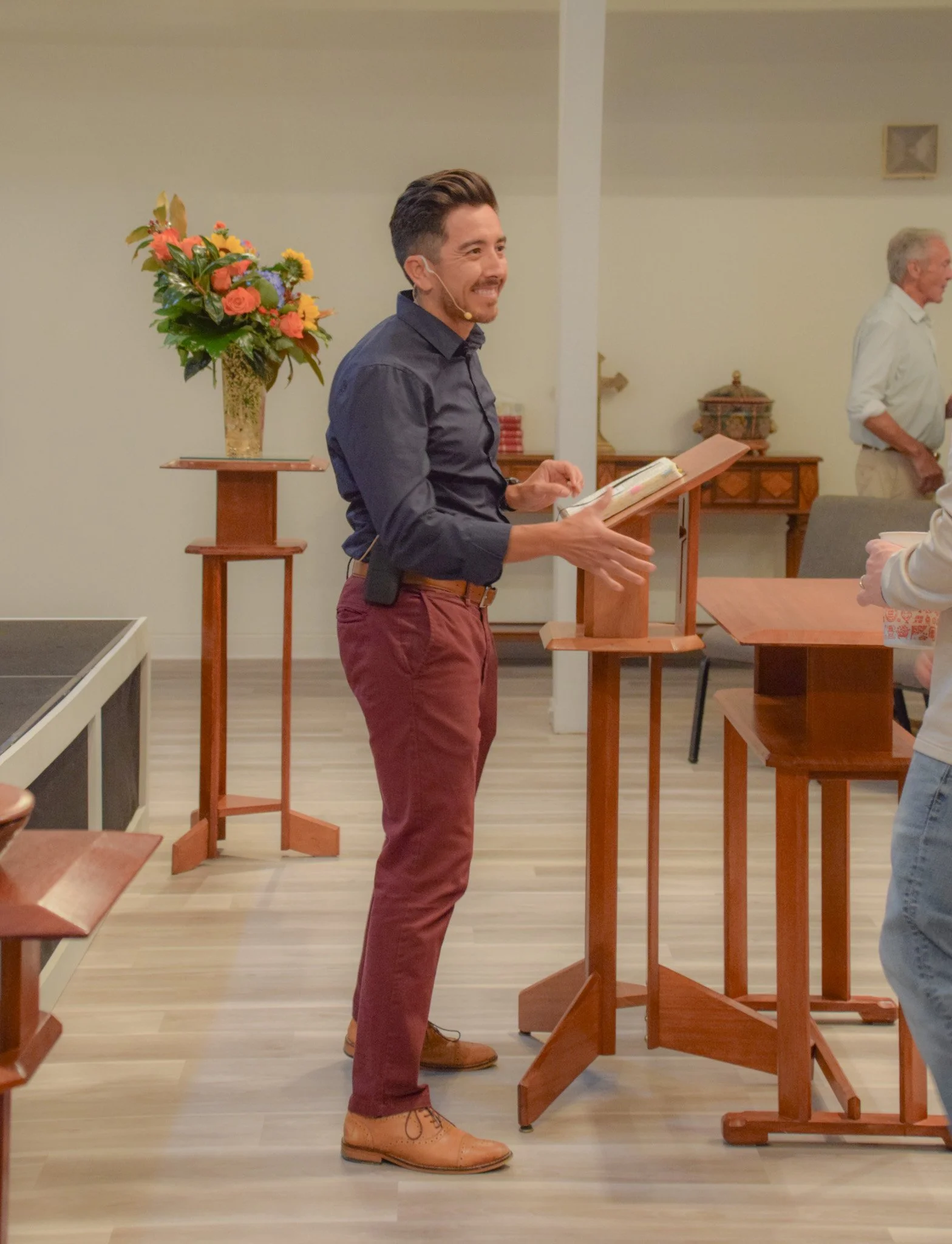 Man with dark hair and beard smiling, wearing a navy blue shirt, maroon pants, and light brown shoes, standing and speaking at a podium in a room decorated with floral arrangements.